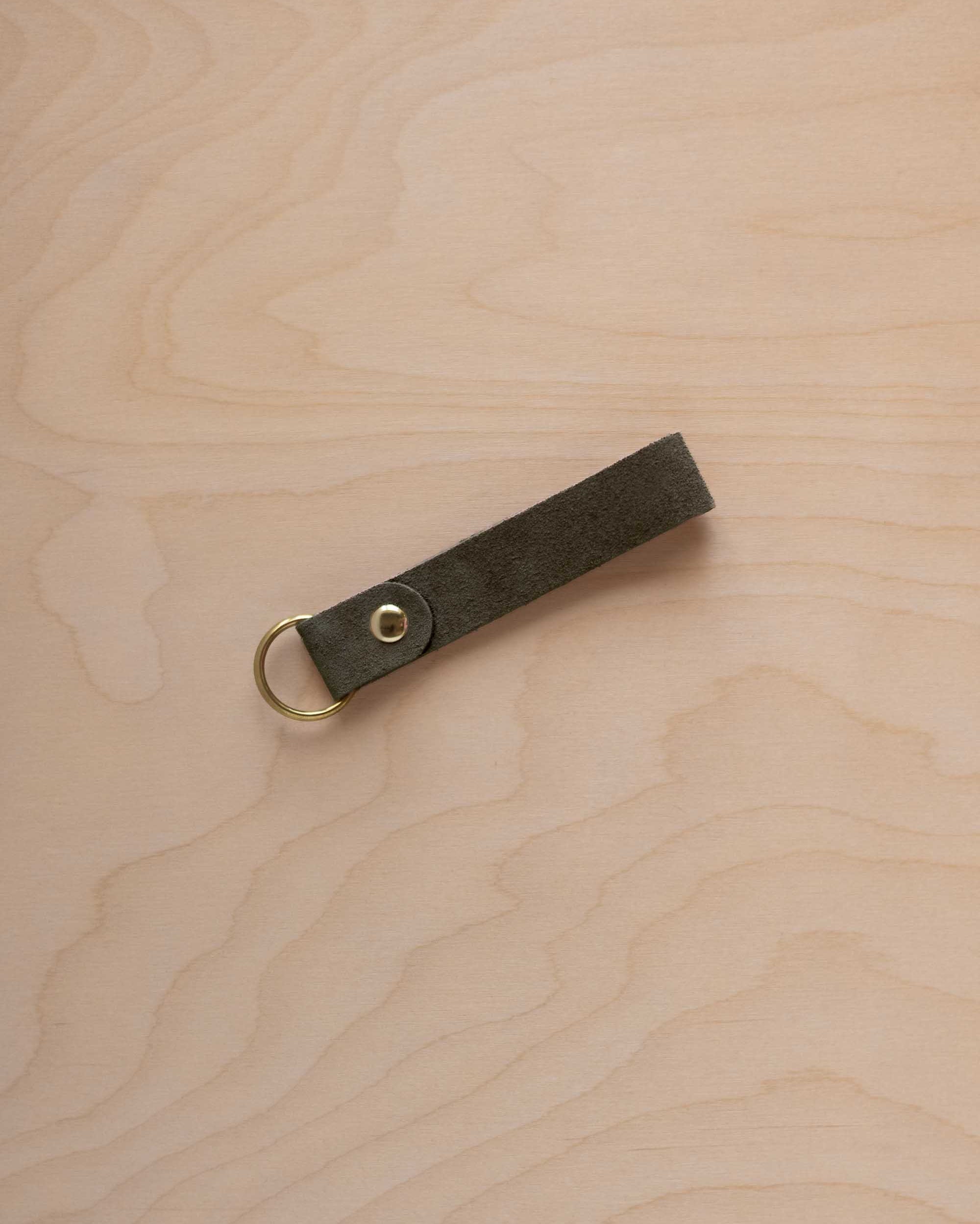 A soft olive green suede leather keychain with a gold metal ring and rivet, displayed diagonally on a light wood grain background.