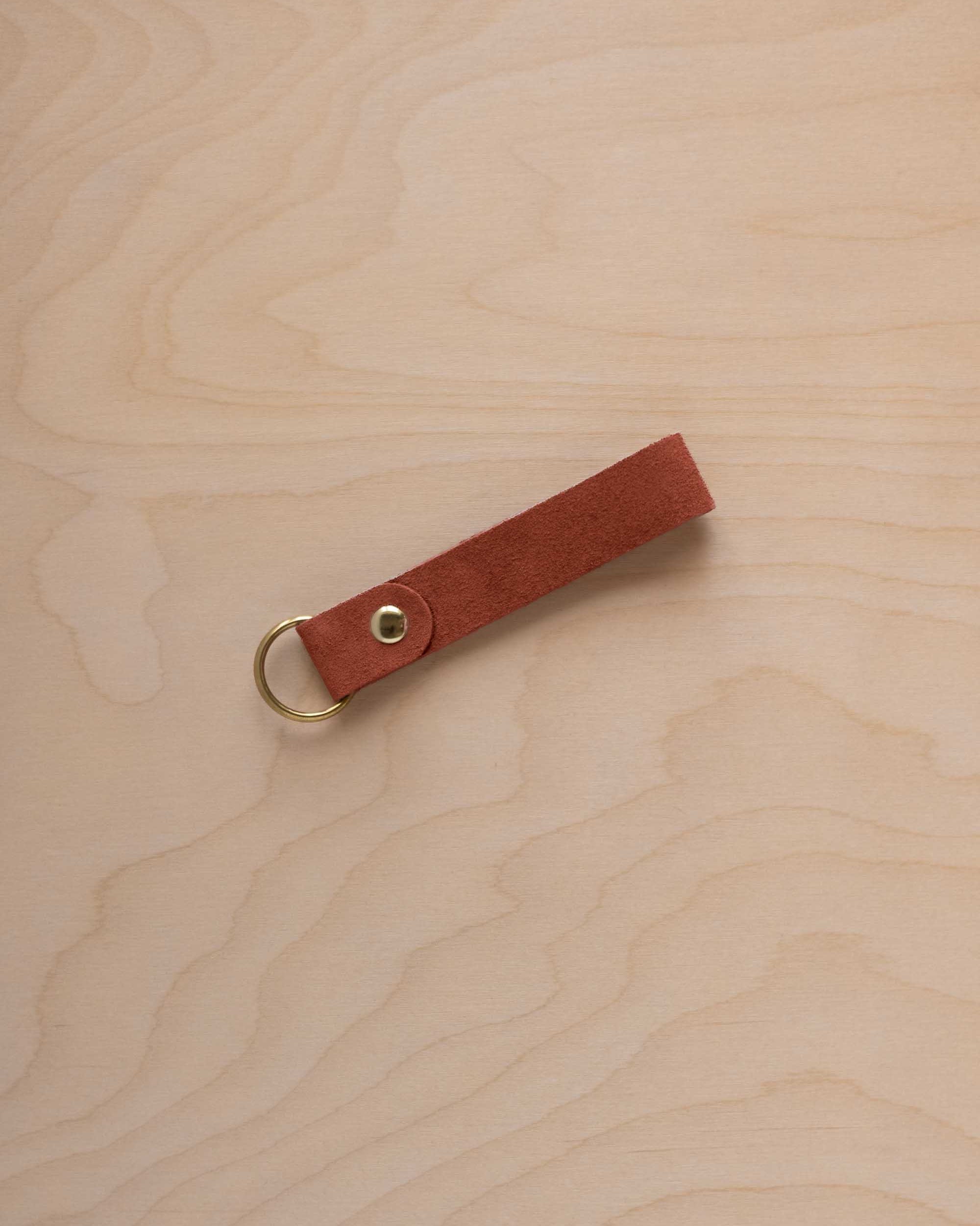 A terracotta suede leather keychain with a gold metal ring and rivet, displayed diagonally on a light wood grain background.
