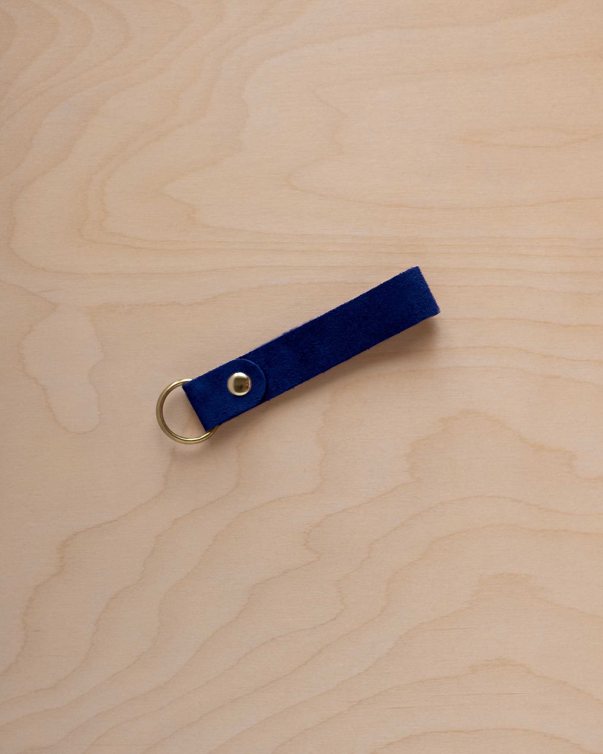 A ultramarine blue suede leather keychain with a gold metal ring and rivet, displayed diagonally on a light wood grain background.