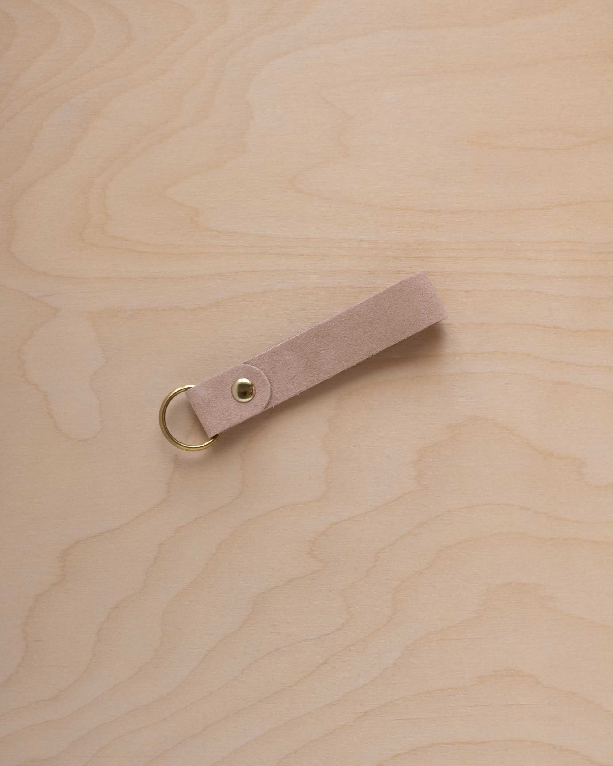 A warm beige suede leather keychain with a gold metal ring and rivet, displayed diagonally on a light wood grain background.