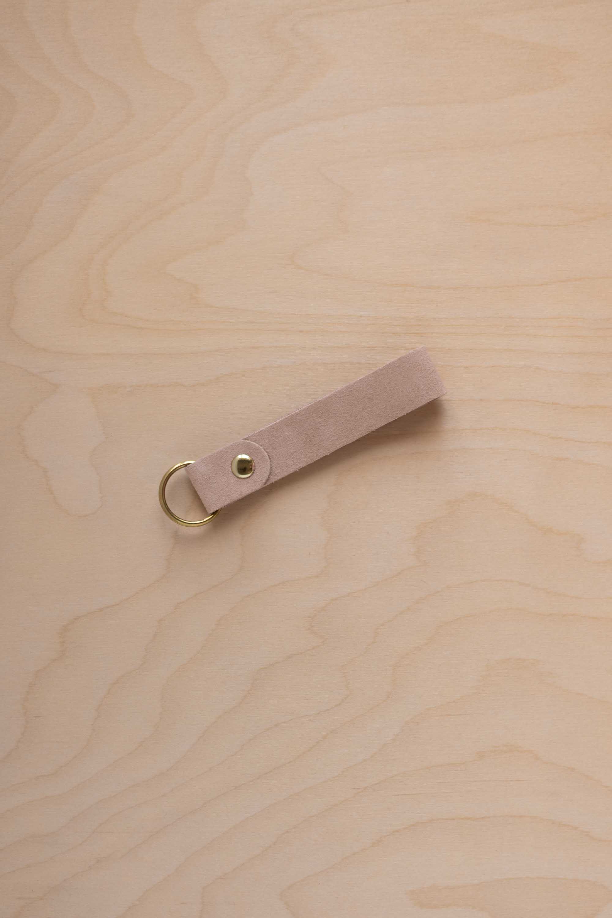 A warm beige suede leather keychain with a gold metal ring and rivet, displayed diagonally on a light wood grain background.