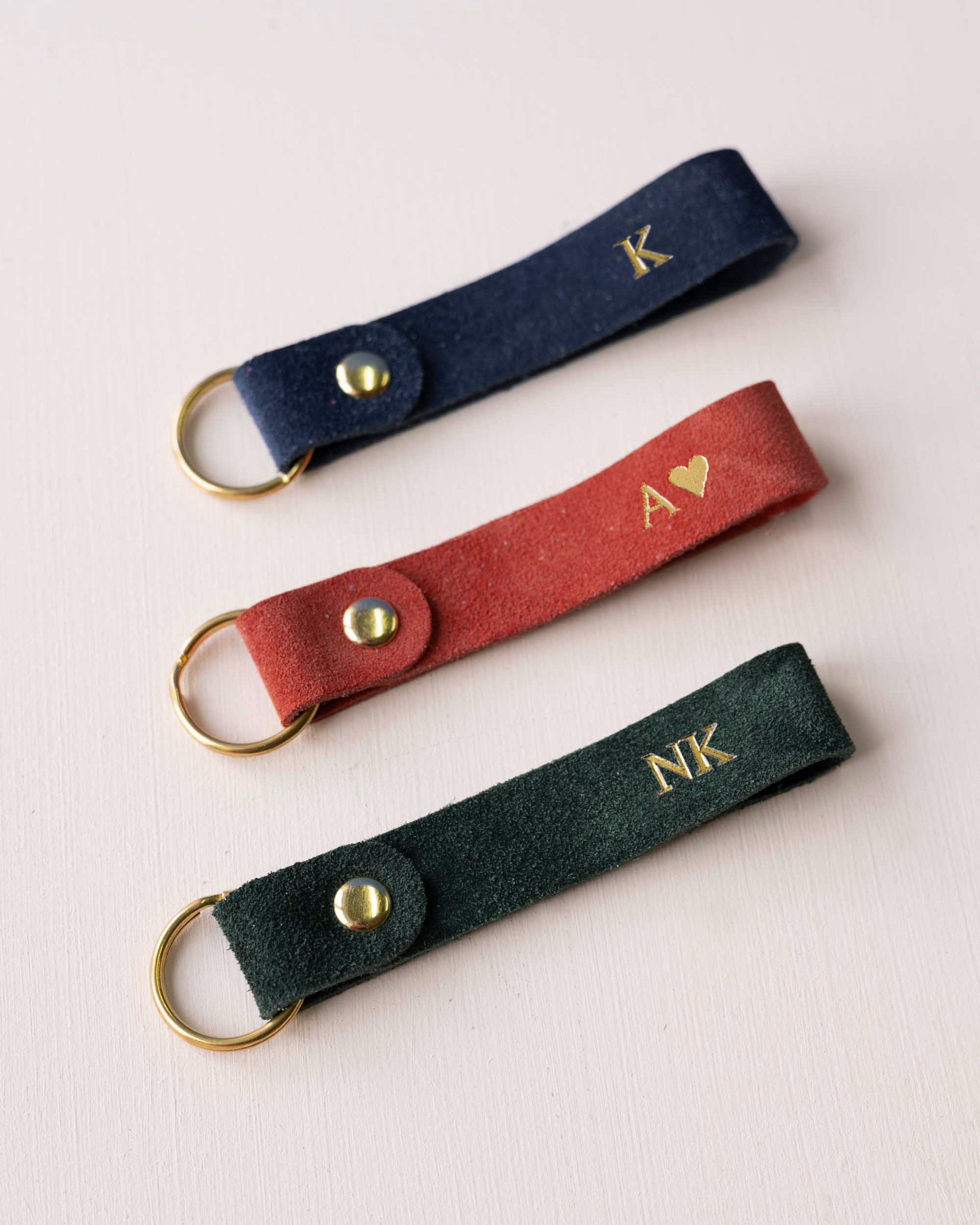 Three personalized suede keychains in navy blue, rust red, and dark green, each featuring a gold-tone metal ring and snap button with custom gold initials or lettering, arranged diagonally on a light background.