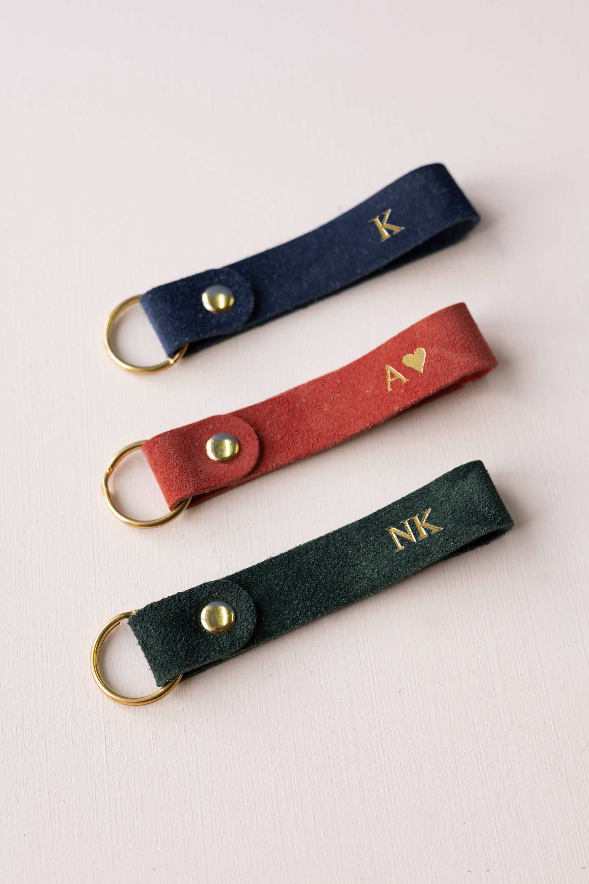 Three personalized suede keychains in navy blue, rust red, and dark green, each featuring a gold-tone metal ring and snap button with custom gold initials or lettering, arranged diagonally on a light background.