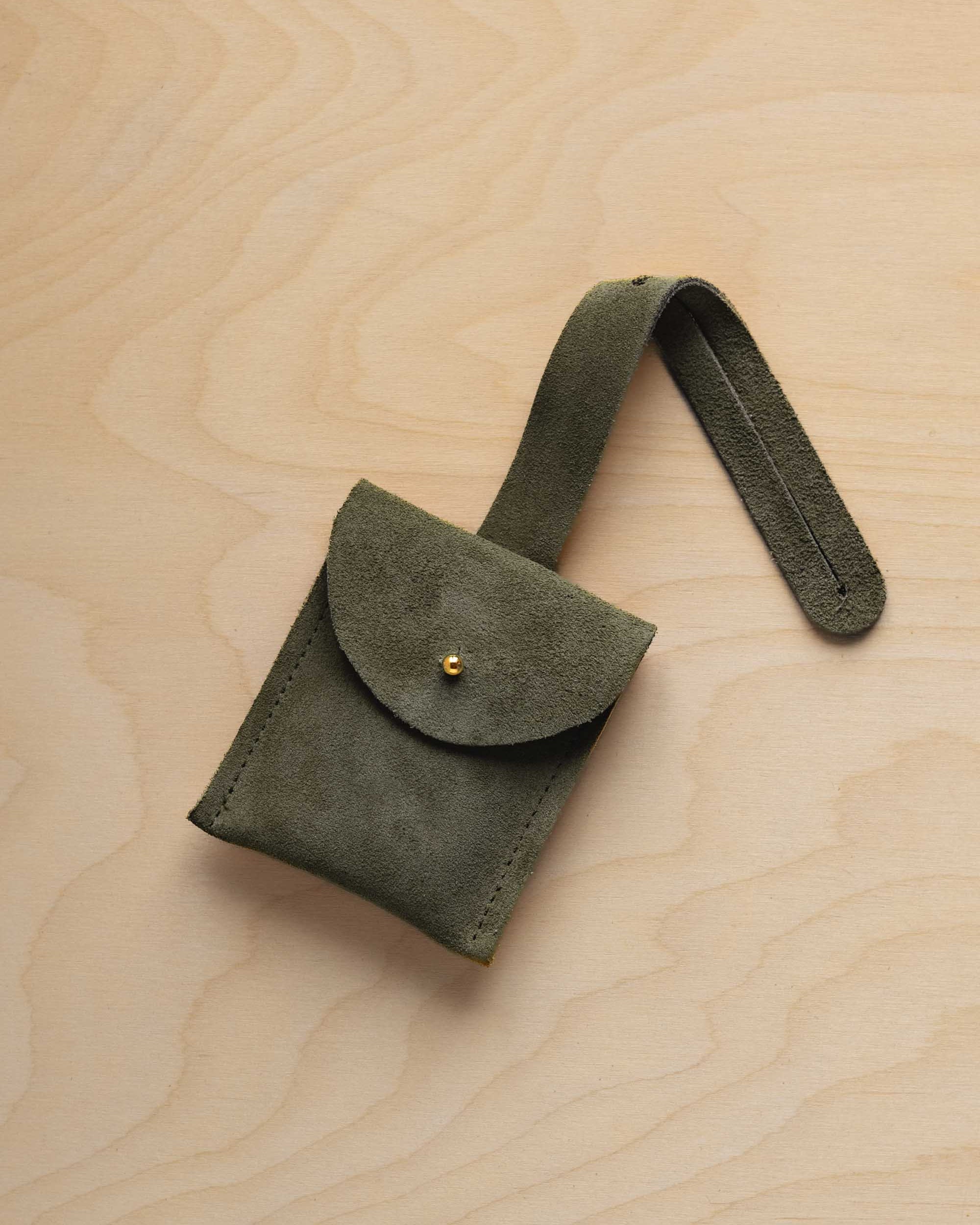A small soft olive green suede pouch with a flap closure and a matching strap, resting on a light wood grain surface.
