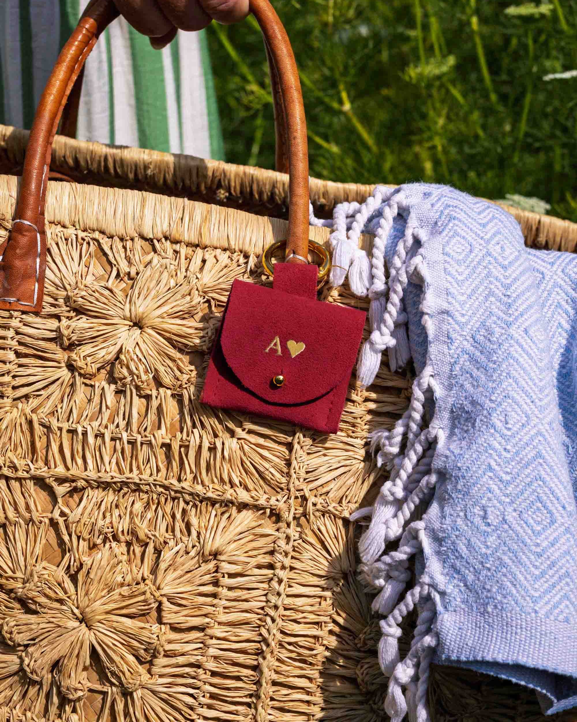 A close-up of a woven straw tote bag with a hand holding the top handle. A small red suede pouch with a gold heart logo and monogram hangs from the bag, and a light blue patterned fringed throw blanket is draped over the side.