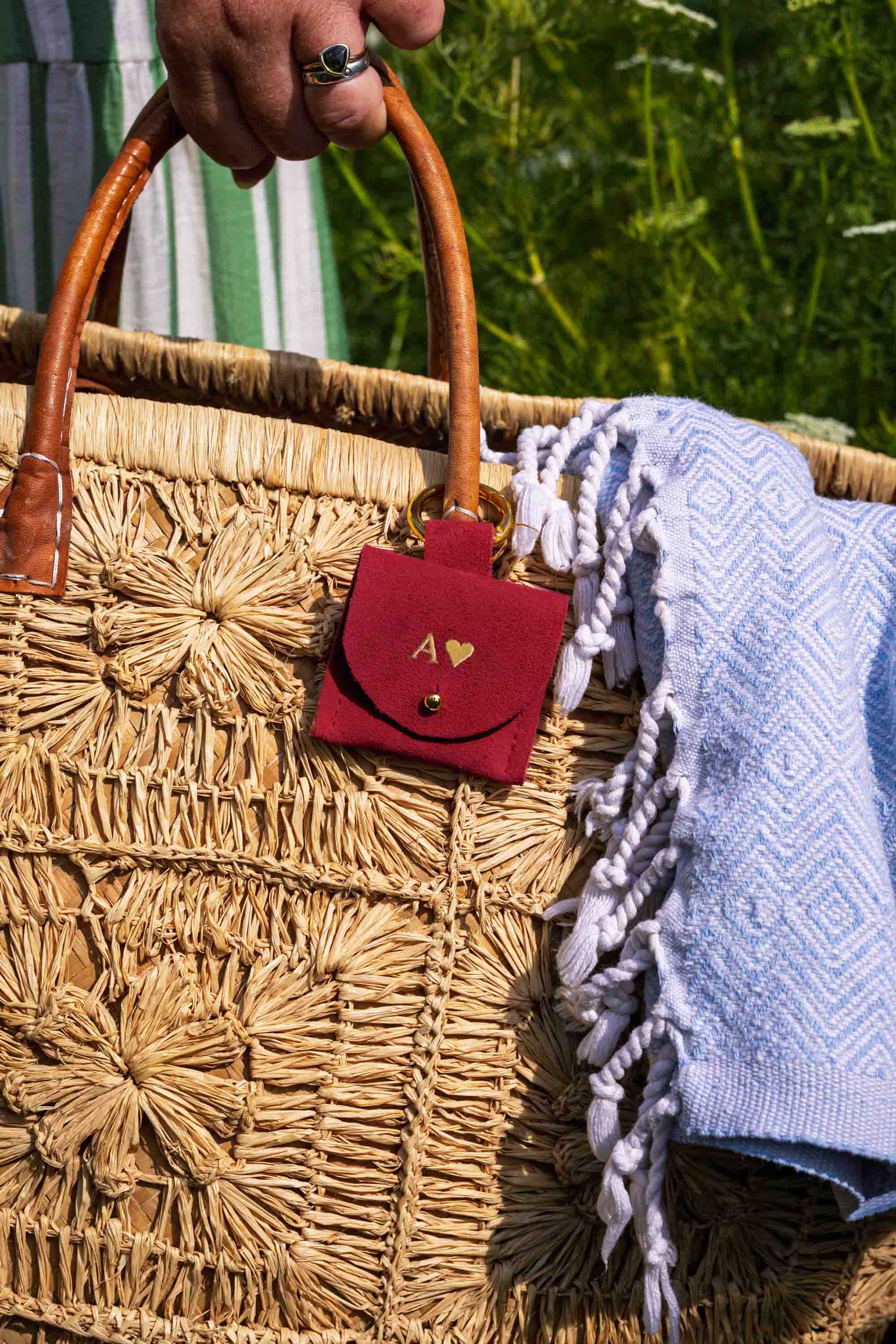 A close-up of a woven straw tote bag with a hand holding the top handle. A small red suede pouch with a gold heart logo and monogram hangs from the bag, and a light blue patterned fringed throw blanket is draped over the side.