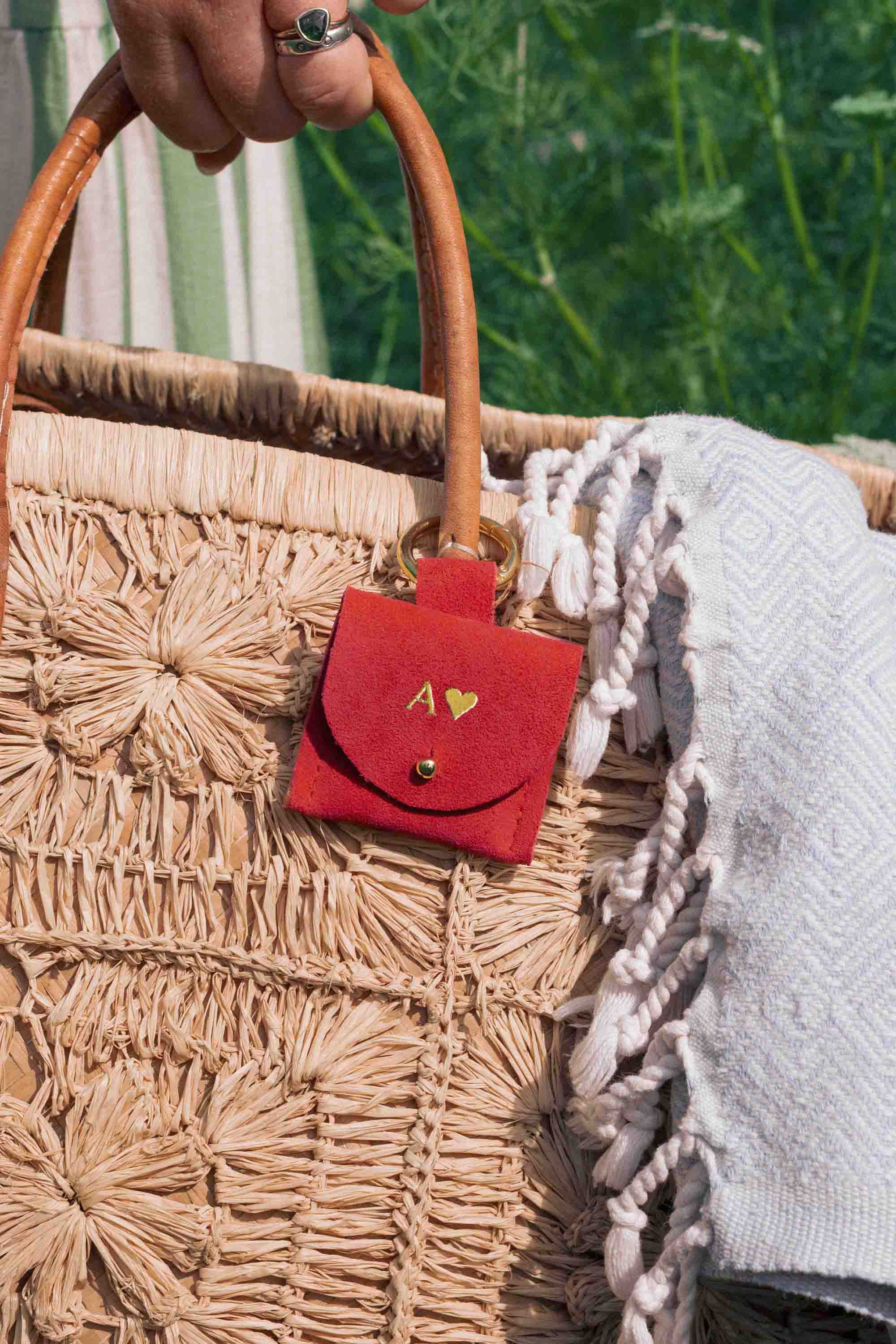 A close-up of a woven straw tote bag with a light brown leather handle, a small red suede pouch charm with gold lettering, and a white beach towel draped over the side.