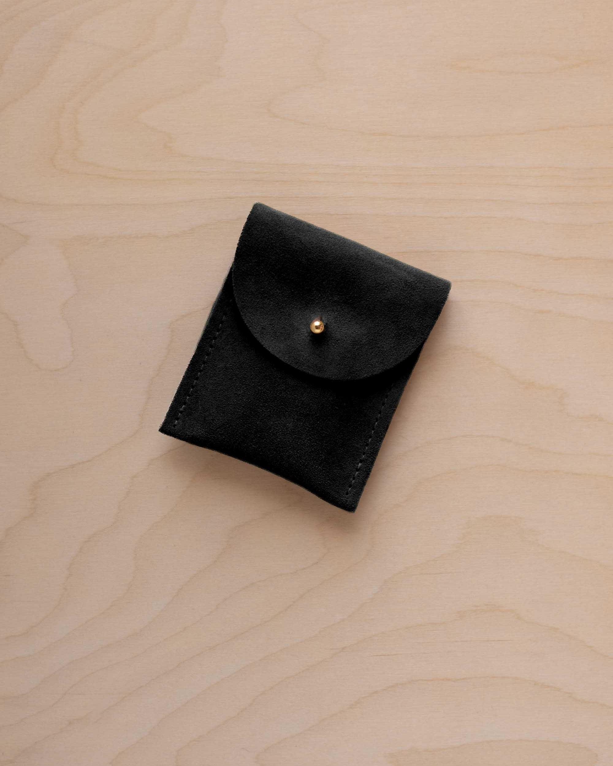 A small black coloured suede pouch with a gold coloured fastening and flap closure, placed on a light wood grain surface.