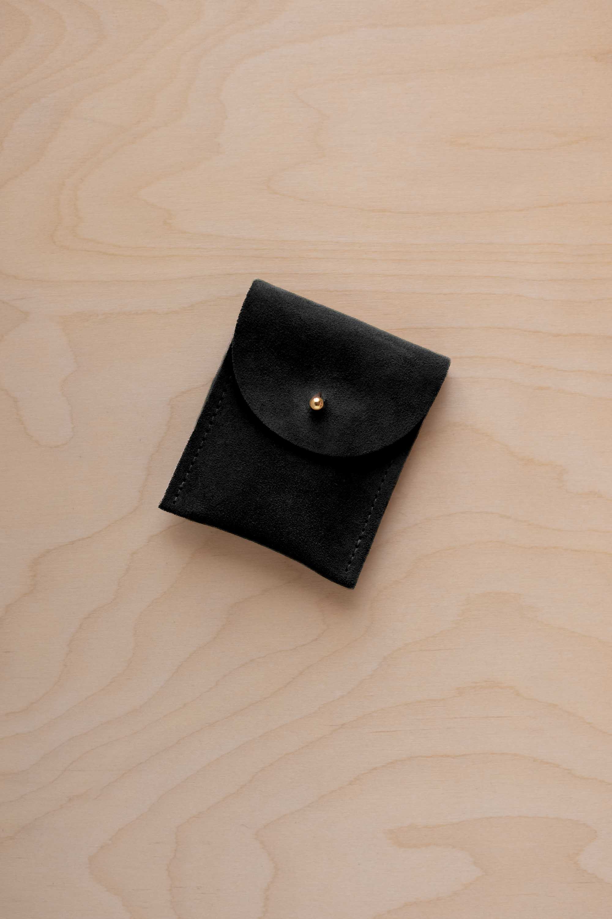 A small black coloured suede pouch with a gold coloured fastening and flap closure, placed on a light wood grain surface.