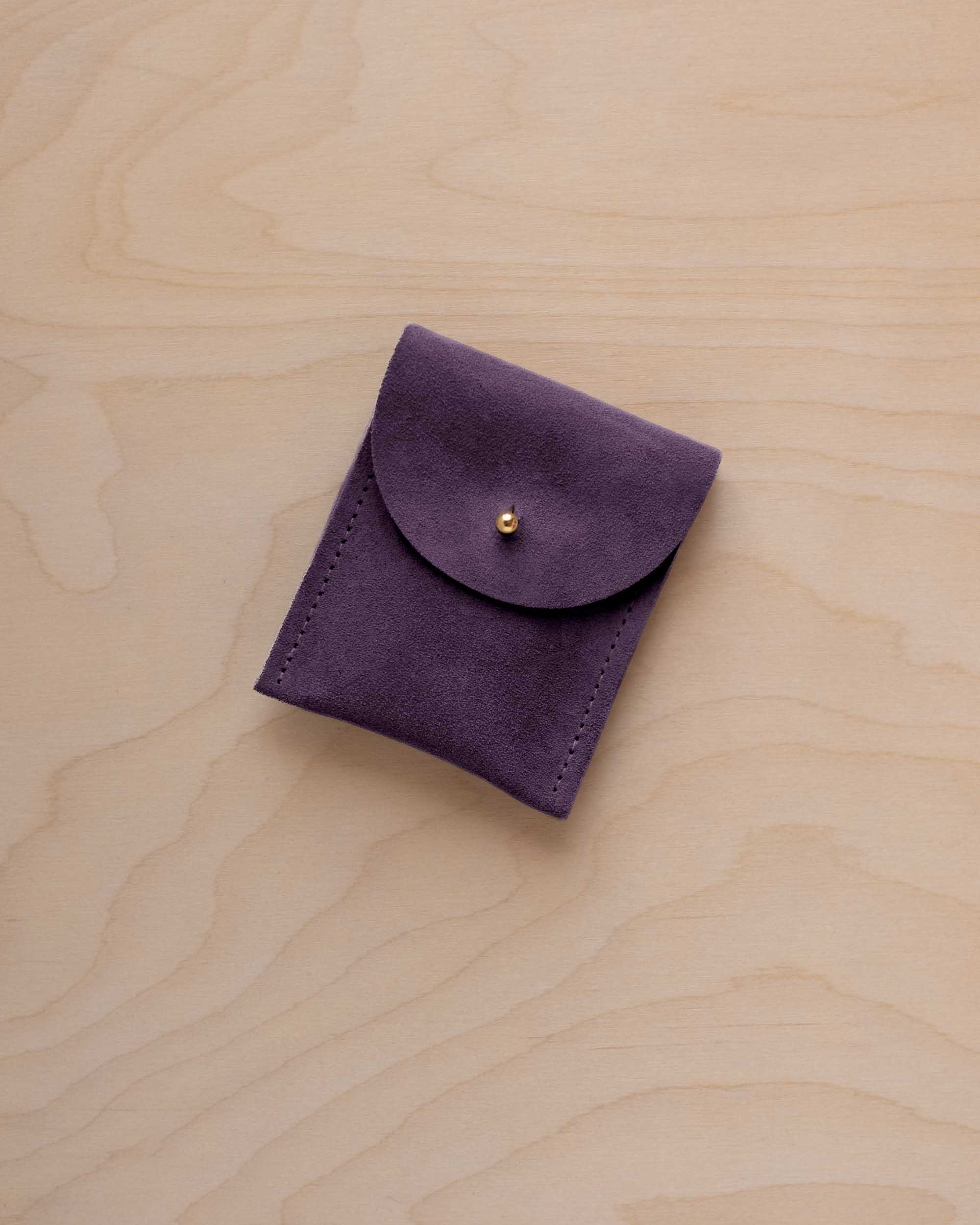 A small blackcurrant coloured suede pouch with a gold coloured fastening and flap closure, placed on a light wood grain surface.