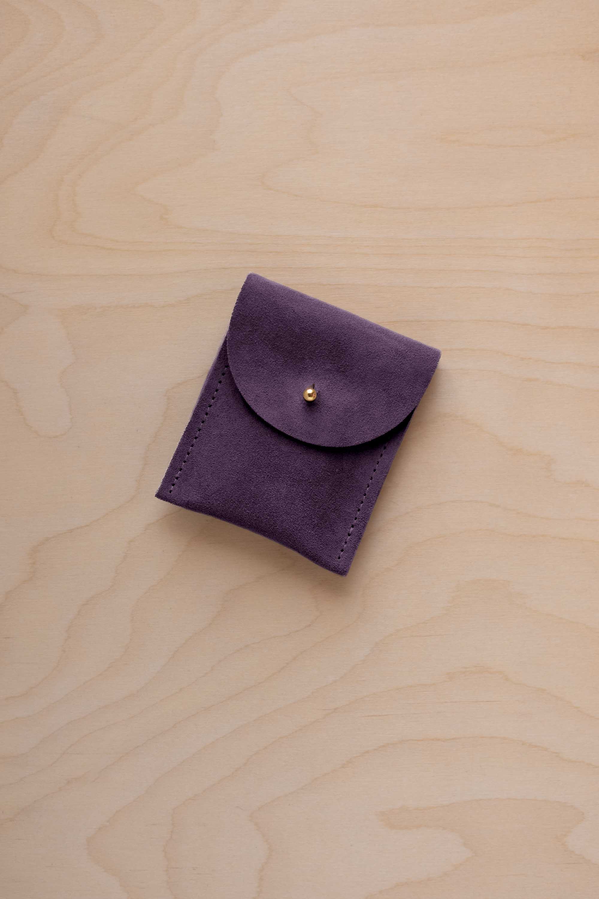A small blackcurrant coloured suede pouch with a gold coloured fastening and flap closure, placed on a light wood grain surface.