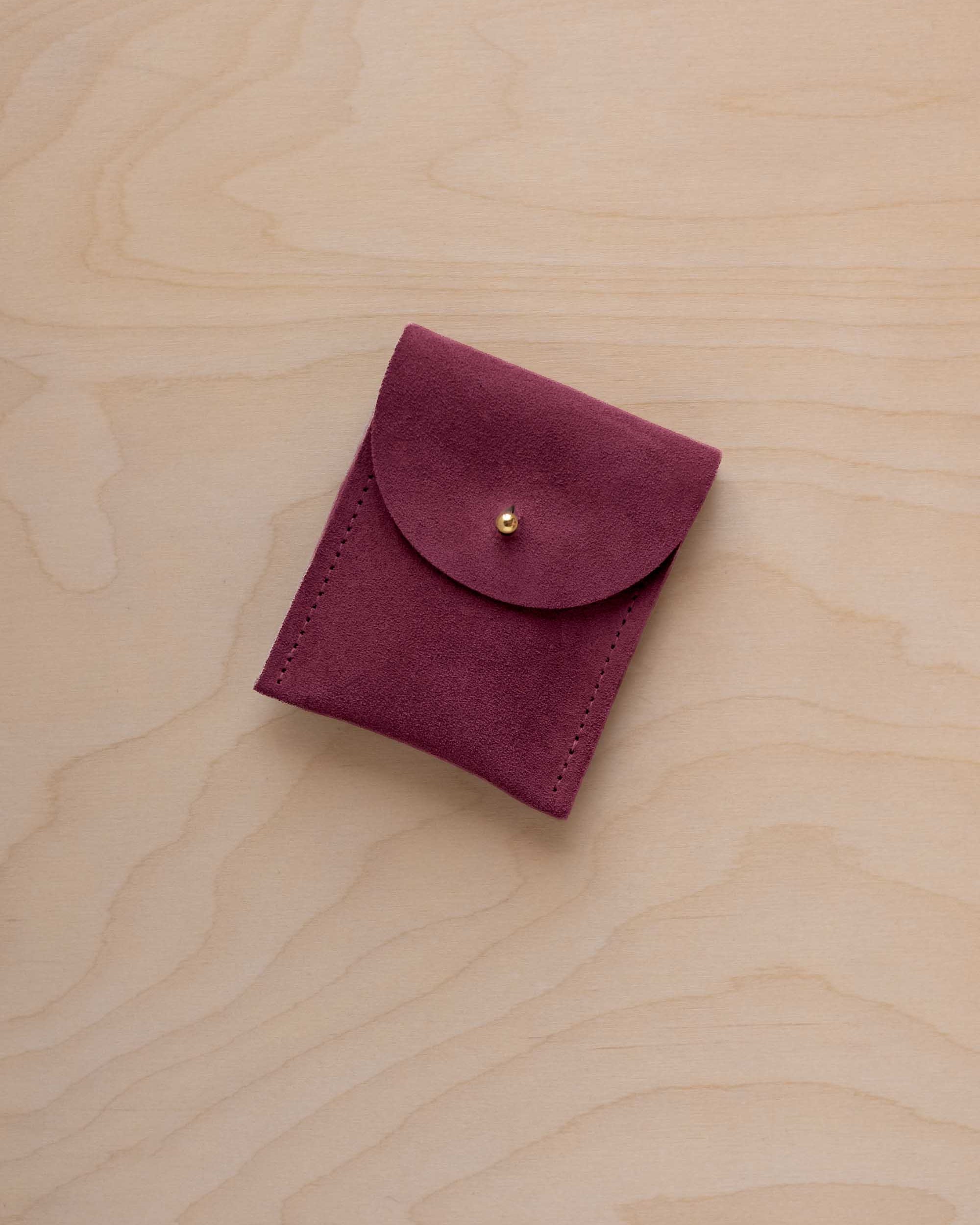 A small burgundy coloured suede pouch with a gold coloured fastening and flap closure, placed on a light wood grain surface.
