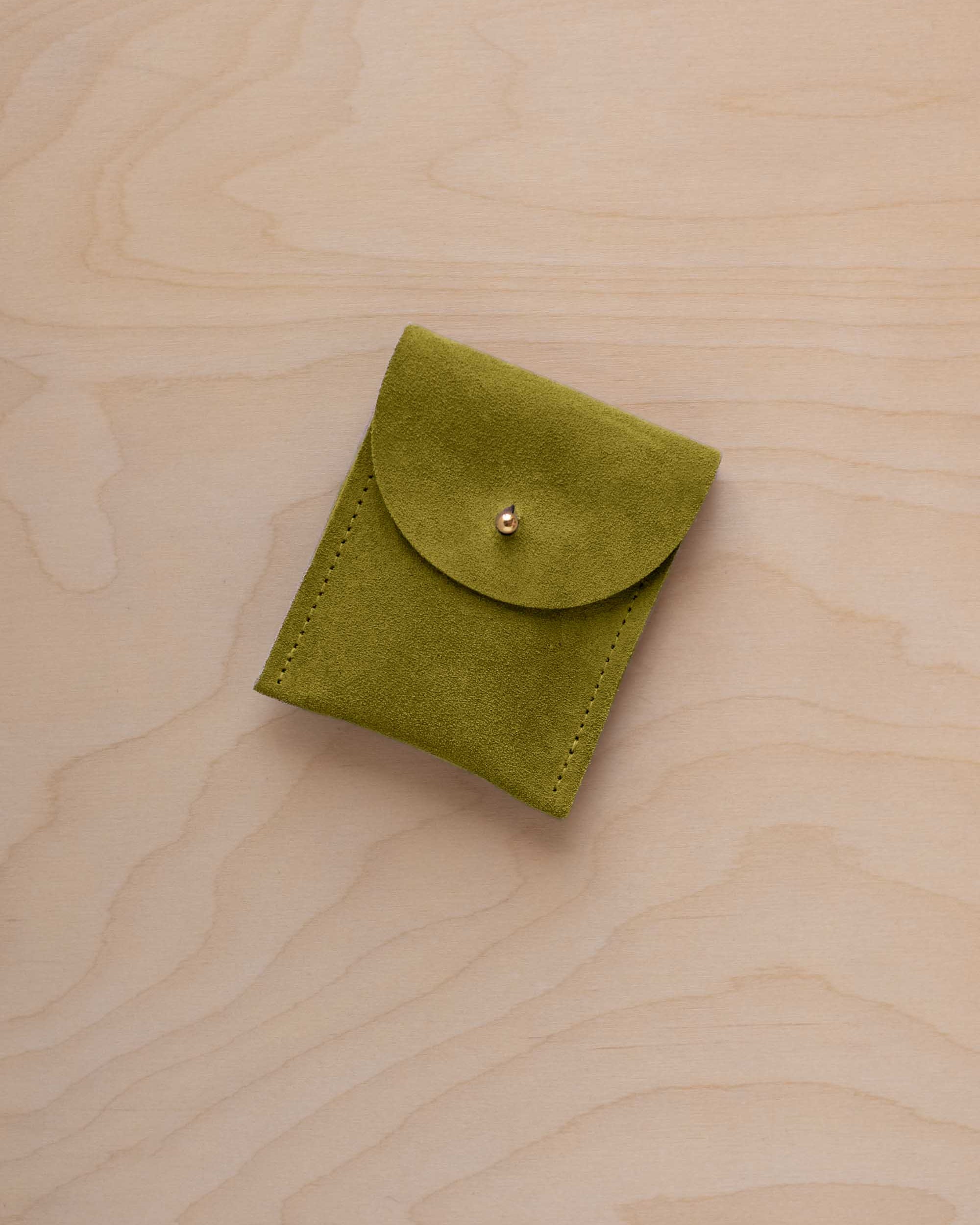 A small chartreuse green coloured suede pouch with a gold coloured fastening and flap closure, placed on a light wood grain surface.