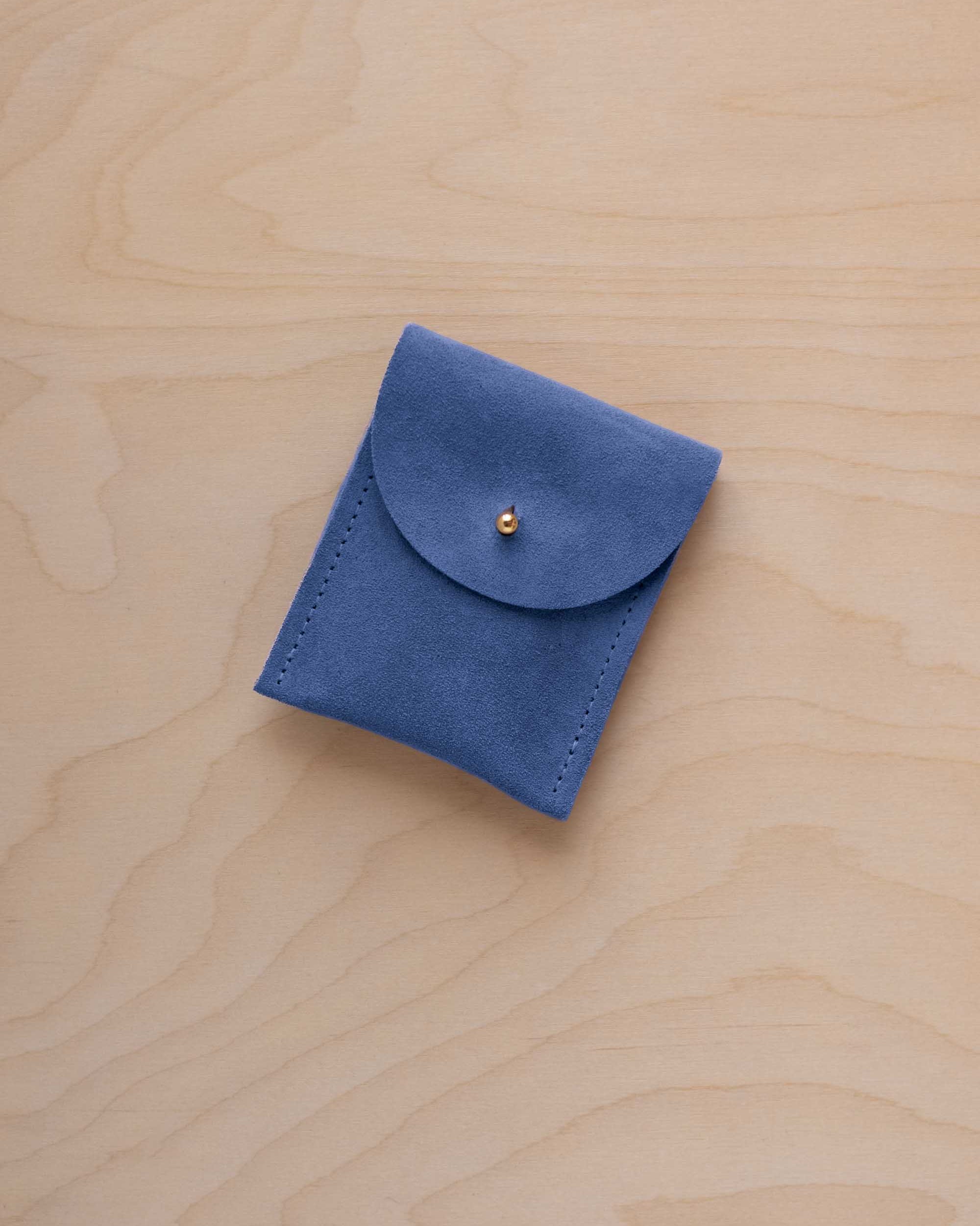 A small cobalt blue coloured suede pouch with a gold coloured fastening and flap closure, placed on a light wood grain surface.