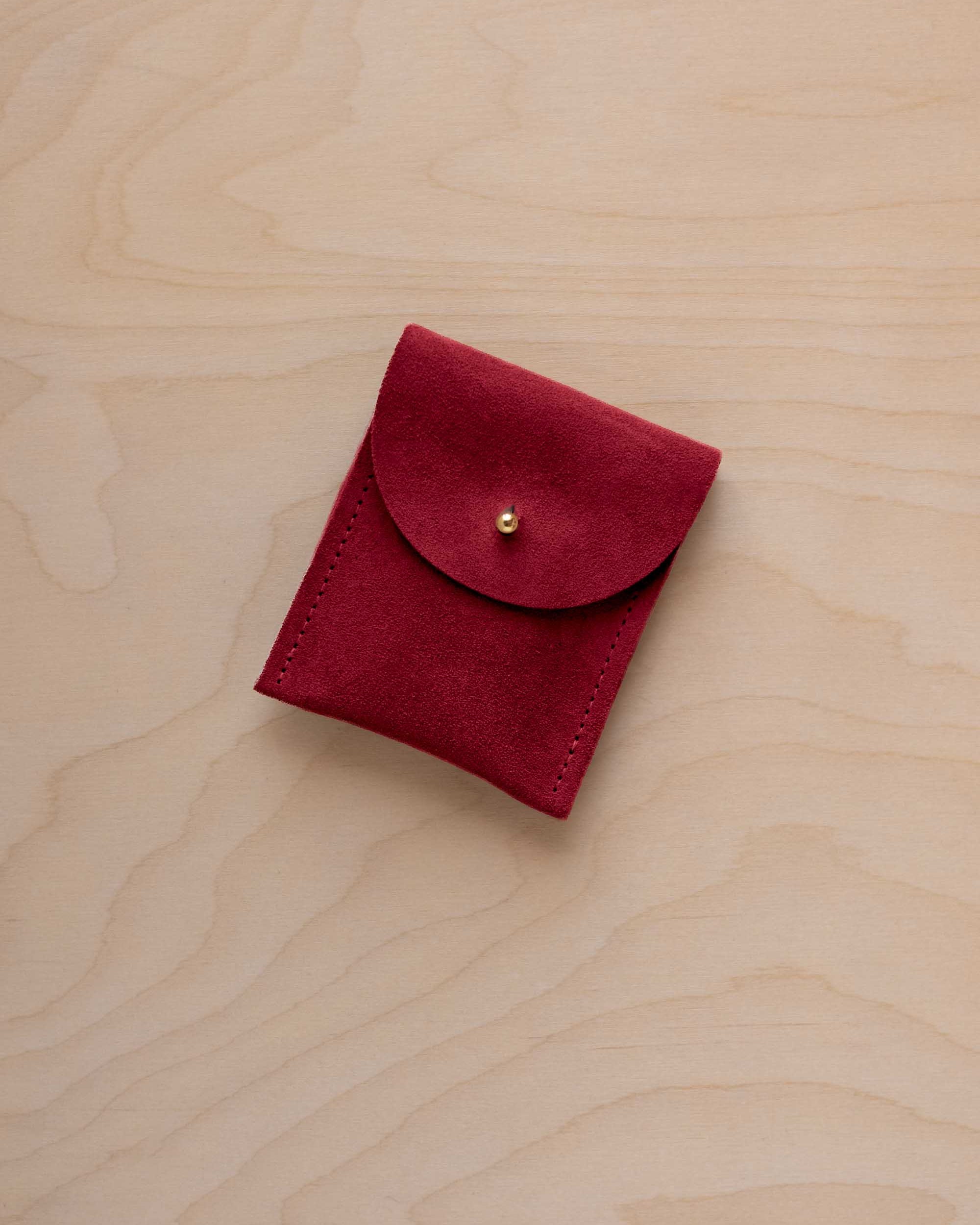 A small crimson red coloured suede pouch with a gold coloured fastening and flap closure, placed on a light wood grain surface.