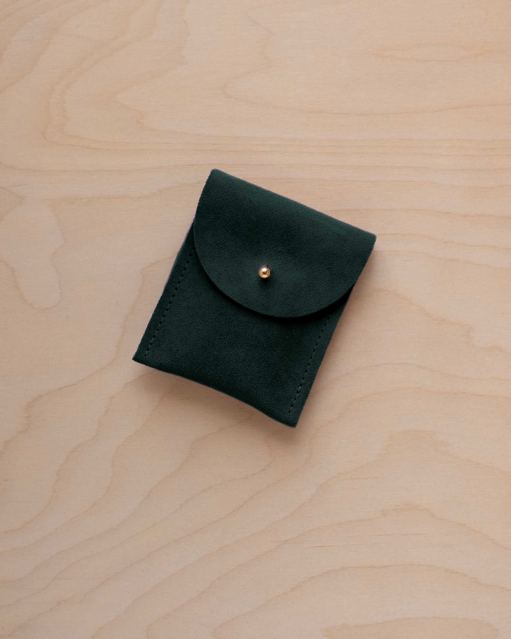 A small deep forest green coloured suede pouch with a gold coloured fastening and flap closure, placed on a light wood grain surface.