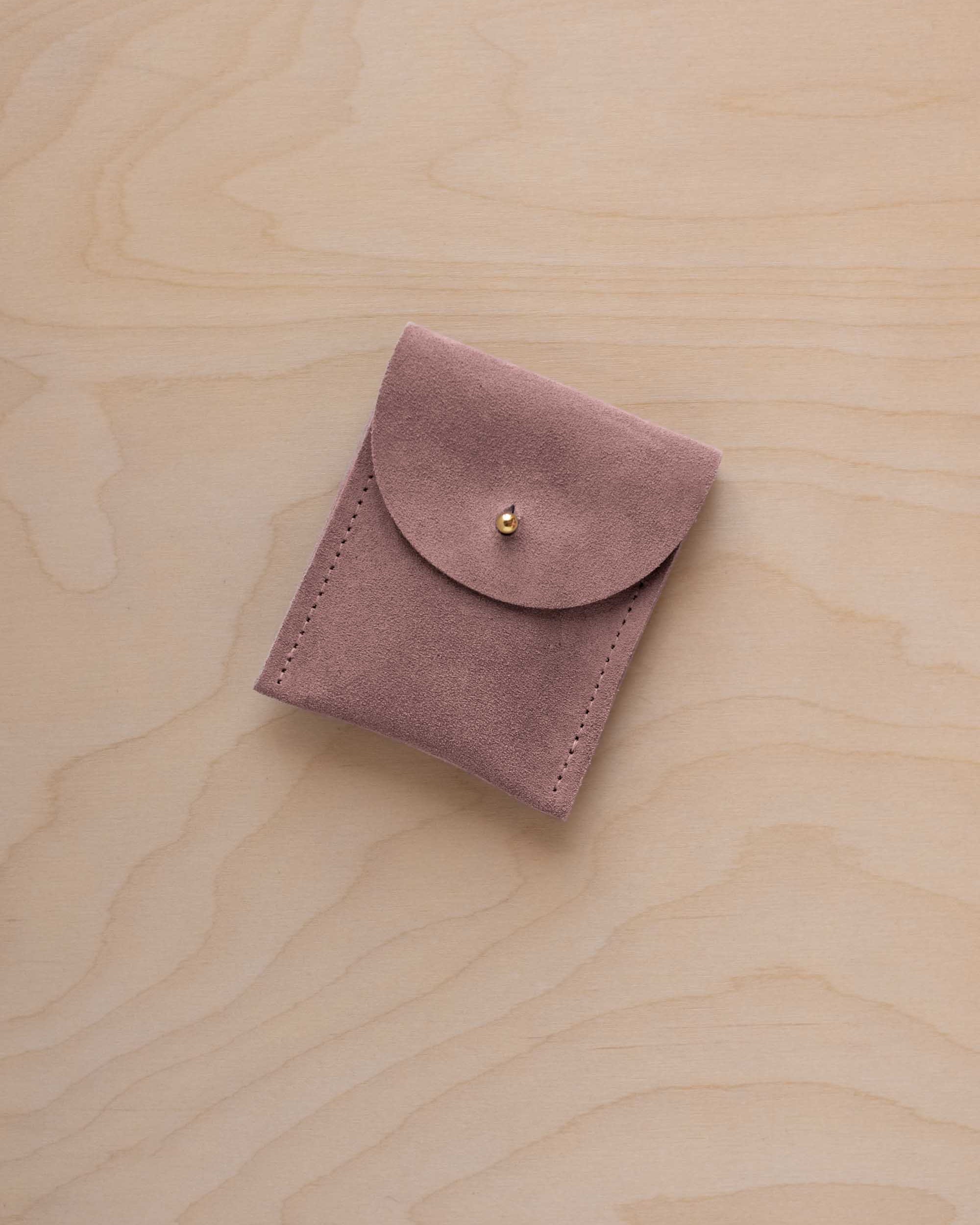 A small dusty mauve coloured suede pouch with a gold coloured fastening and flap closure, placed on a light wood grain surface.