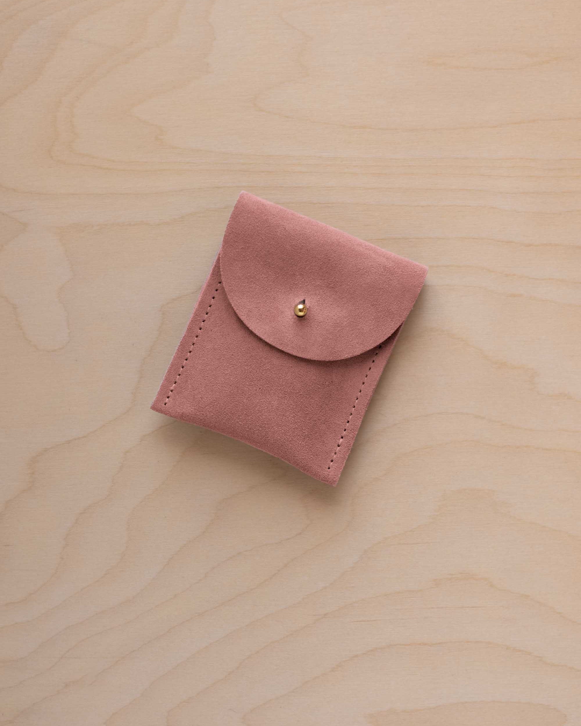 A small dusty rose coloured suede pouch with a gold coloured fastening and flap closure, placed on a light wood grain surface.
