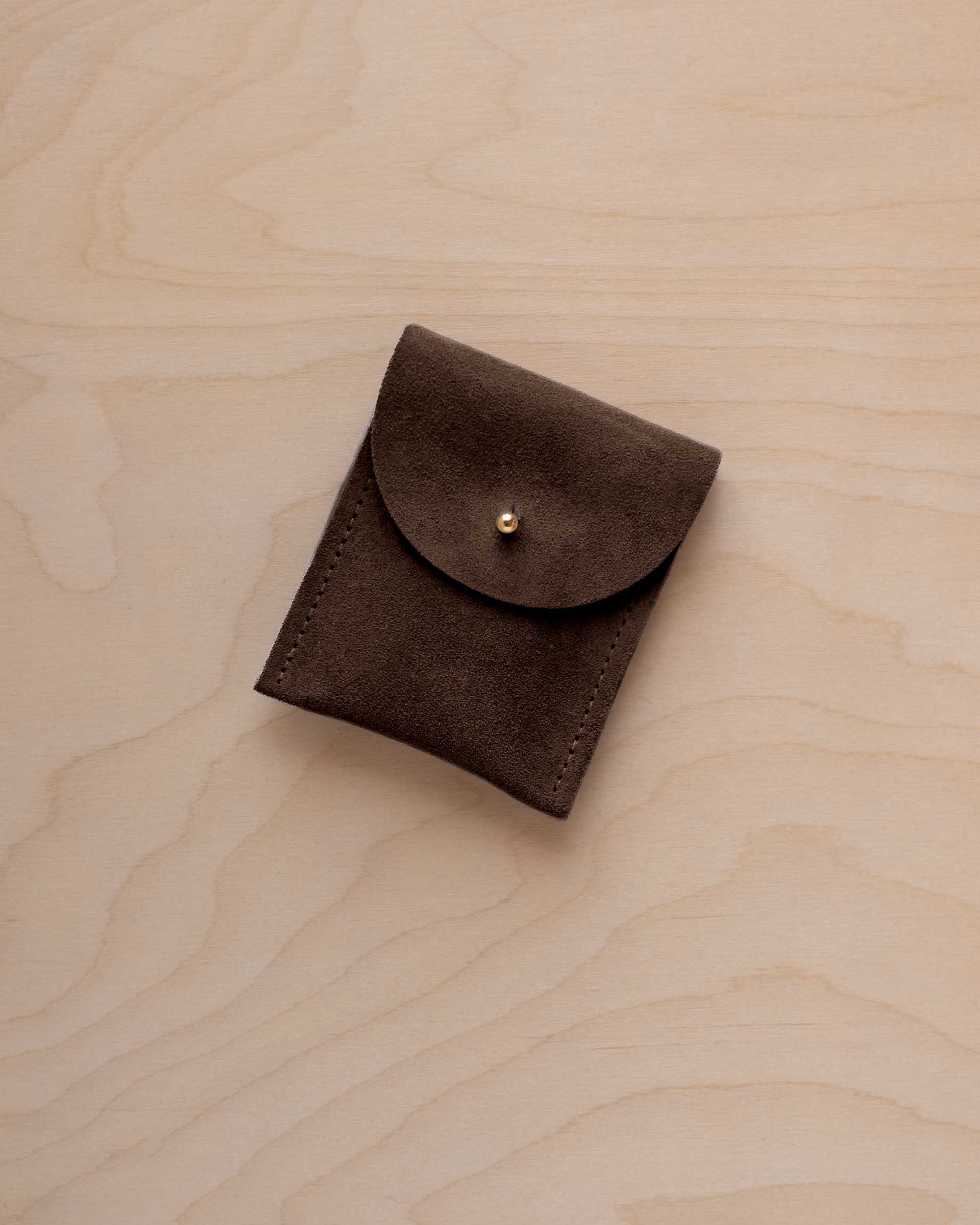 A small espresso brown coloured suede pouch with a gold coloured fastening and flap closure, placed on a light wood grain surface.