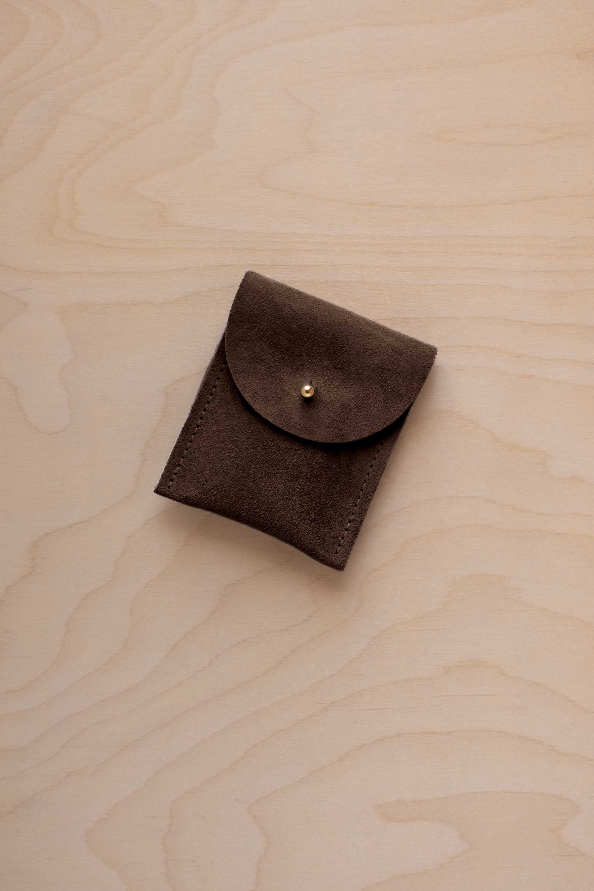 A small espresso brown coloured suede pouch with a gold coloured fastening and flap closure, placed on a light wood grain surface.