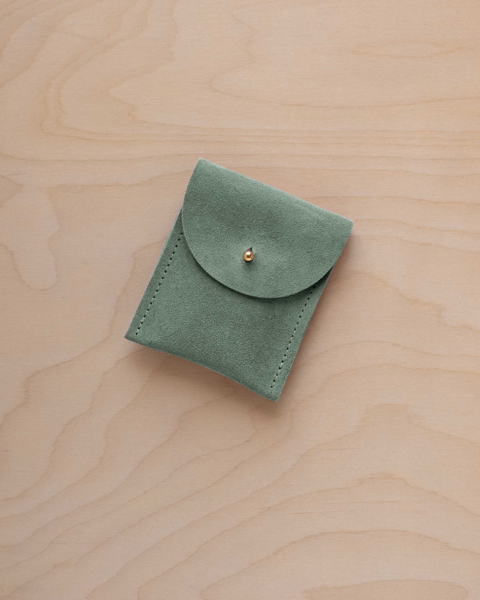 A small eucalyptus green coloured suede pouch with a gold coloured fastening and flap closure, placed on a light wood grain surface.