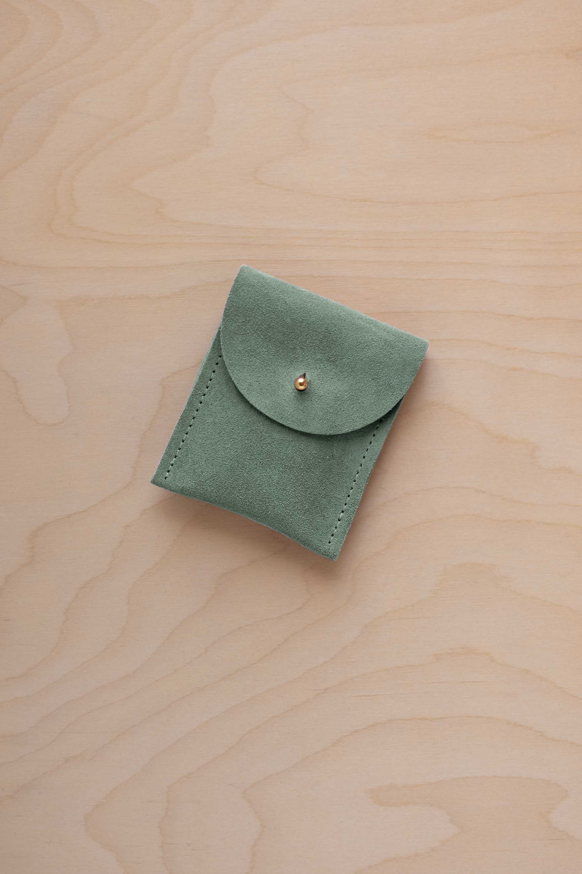 A small eucalyptus green coloured suede pouch with a gold coloured fastening and flap closure, placed on a light wood grain surface.