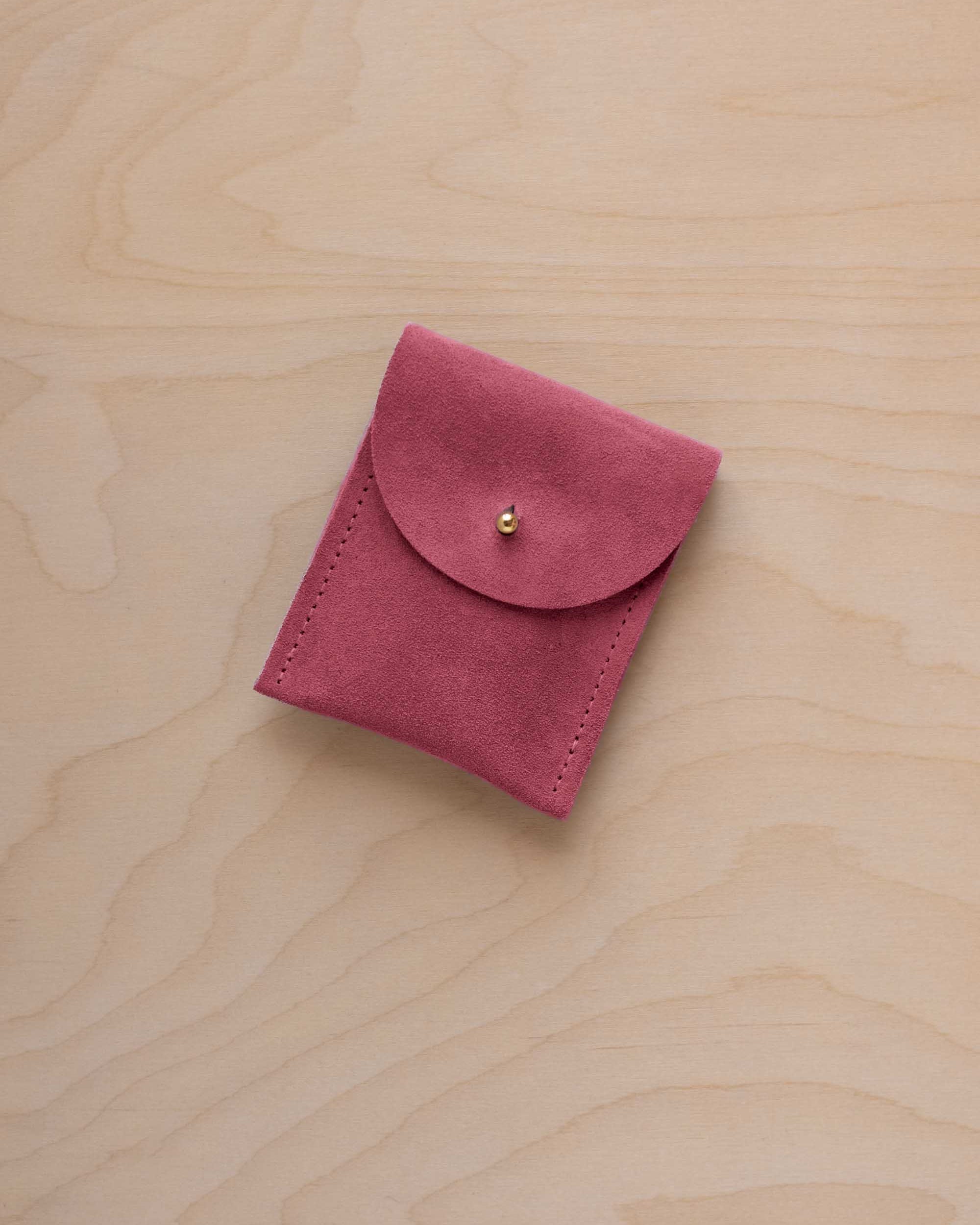 A small faded rose coloured suede pouch with a gold coloured fastening and flap closure, placed on a light wood grain surface.