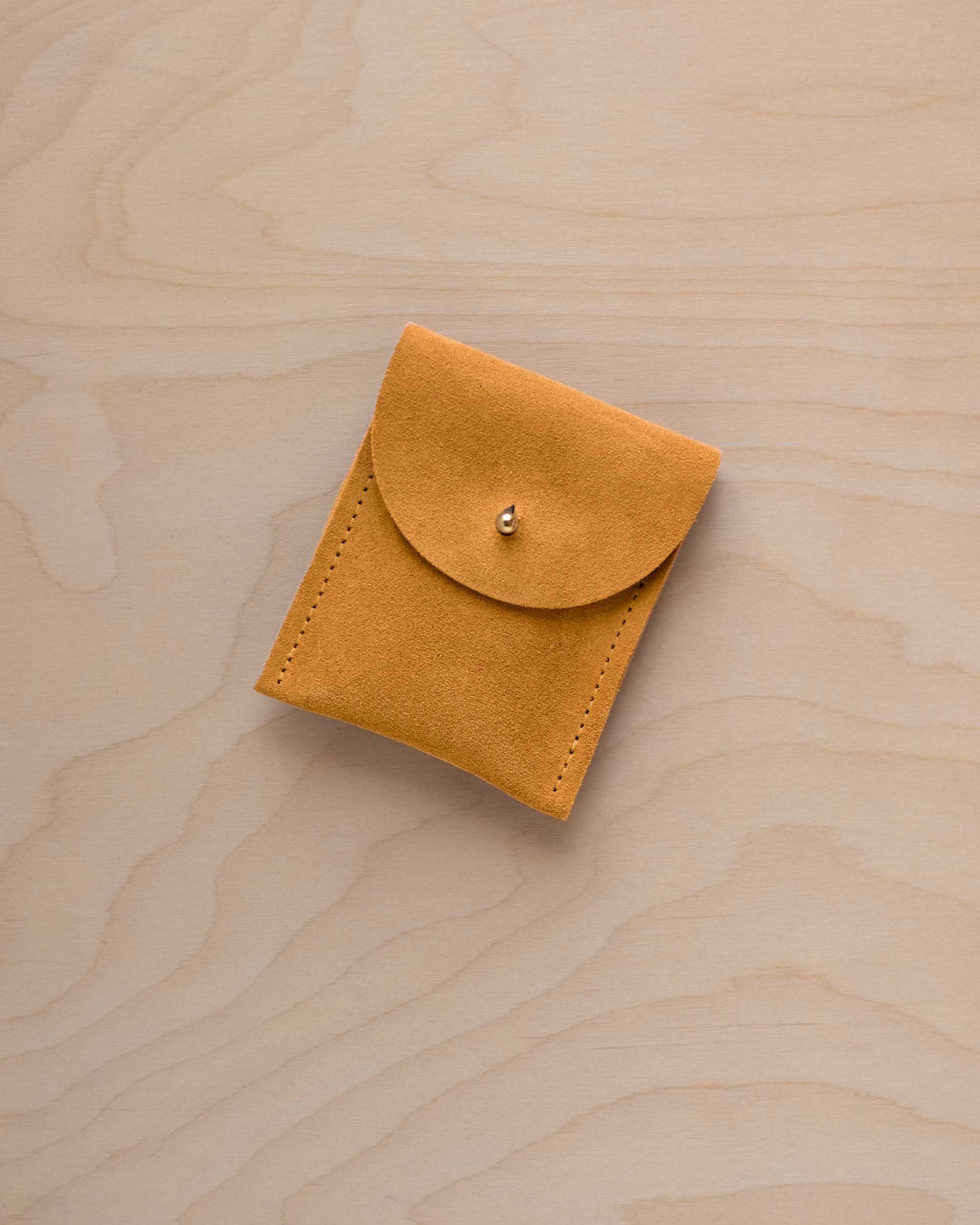 A small golden ochre coloured suede pouch with a gold coloured fastening and flap closure, placed on a light wood grain surface.
