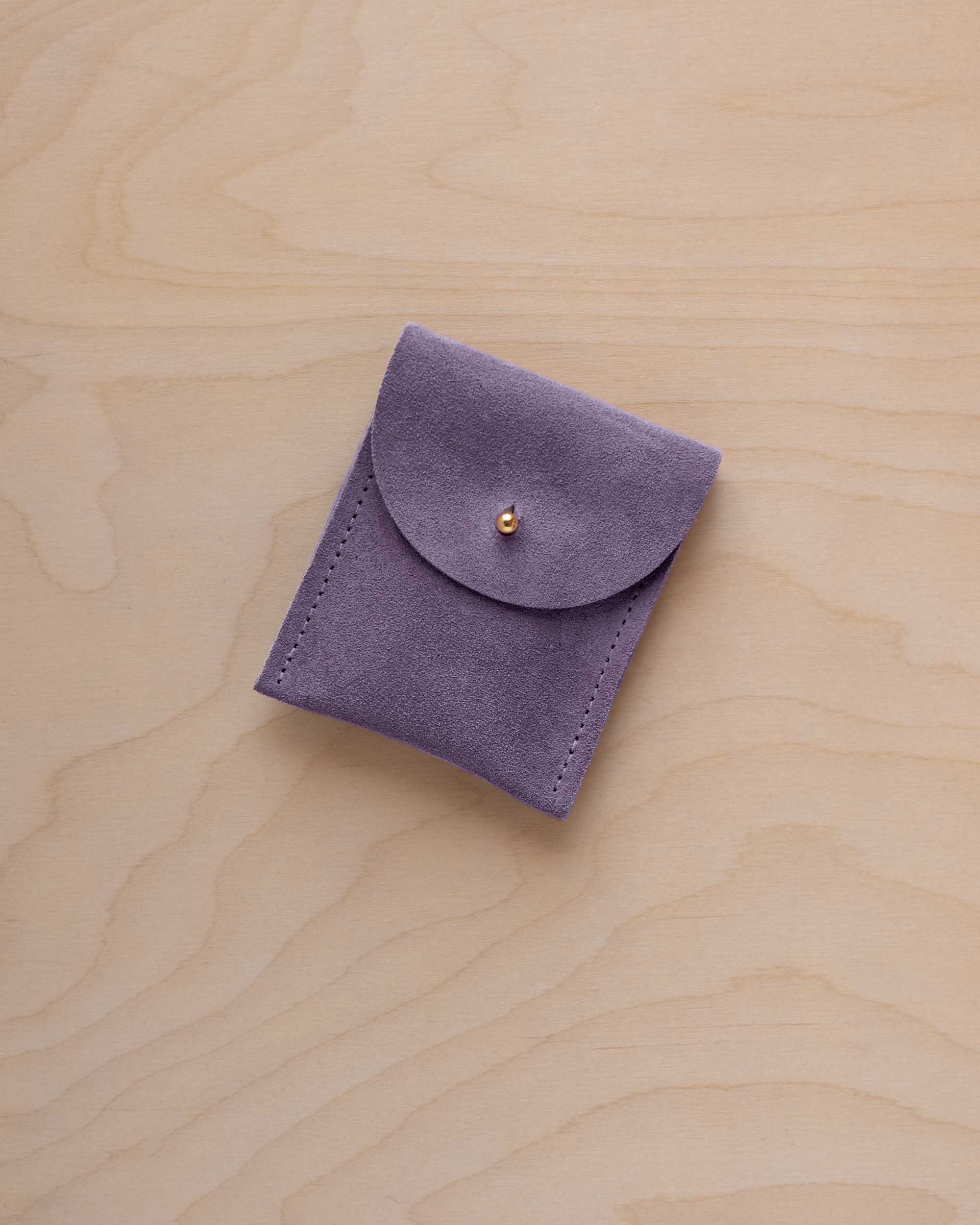 A small lavender coloured suede pouch with a gold coloured fastening and flap closure, placed on a light wood grain surface.