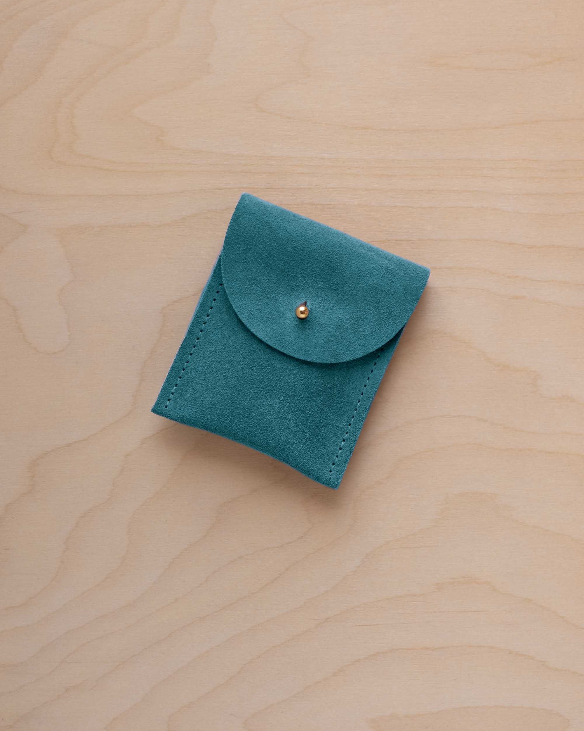 A small marine teal coloured suede pouch with a gold coloured fastening and flap closure, placed on a light wood grain surface.