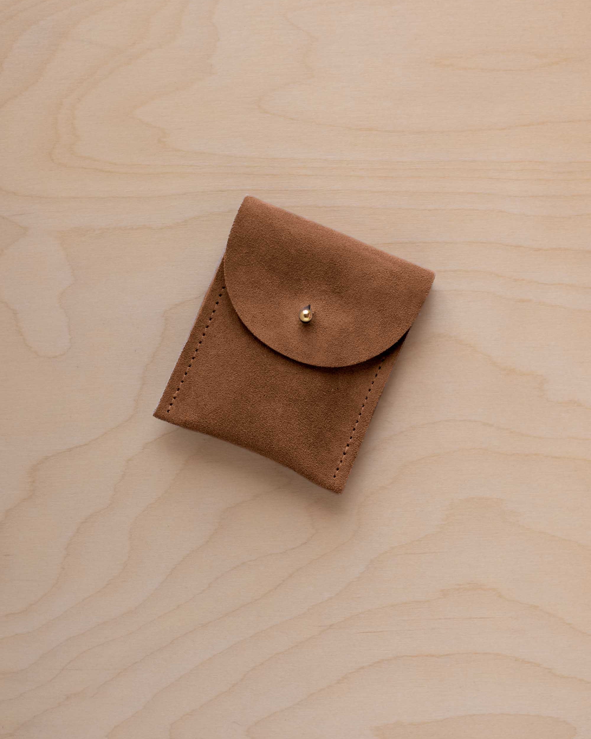 A small mocha brown coloured suede pouch with a gold coloured fastening and flap closure, placed on a light wood grain surface.