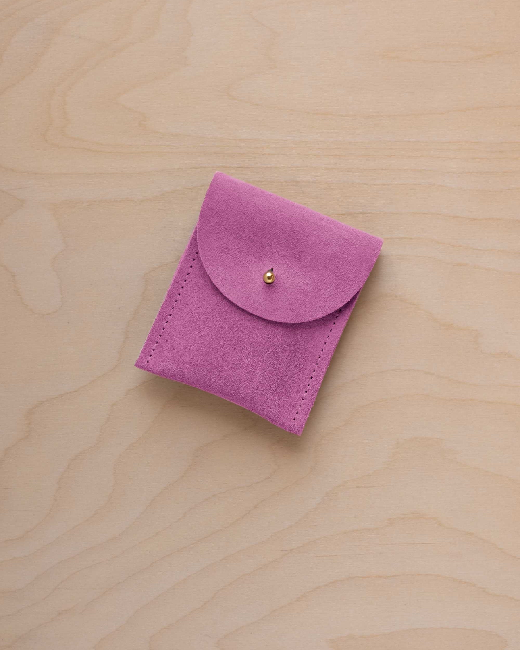 A small peony pink coloured suede pouch with a gold coloured fastening and flap closure, placed on a light wood grain surface.