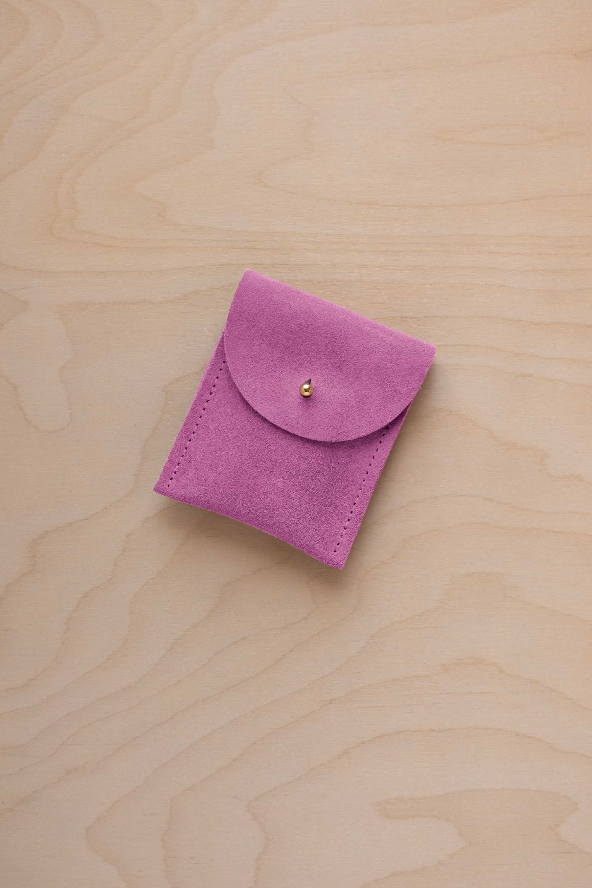 A small peony pink coloured suede pouch with a gold coloured fastening and flap closure, placed on a light wood grain surface.