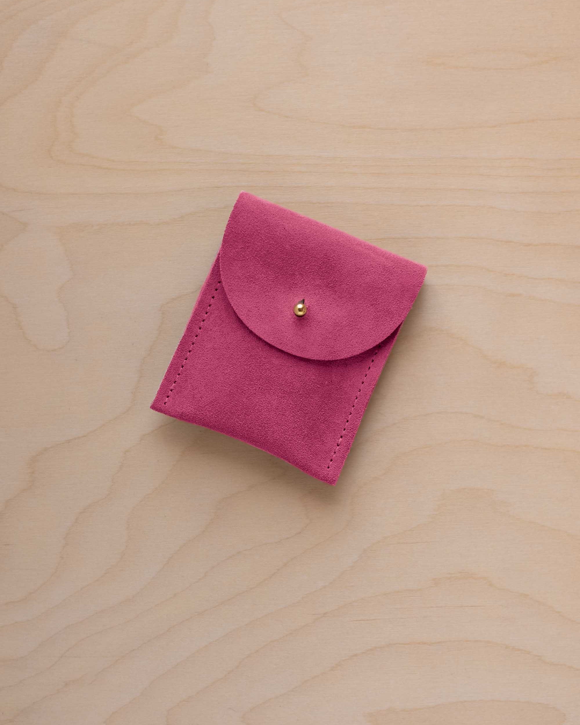 A small raspberry coloured suede pouch with a gold coloured fastening and flap closure, placed on a light wood grain surface.