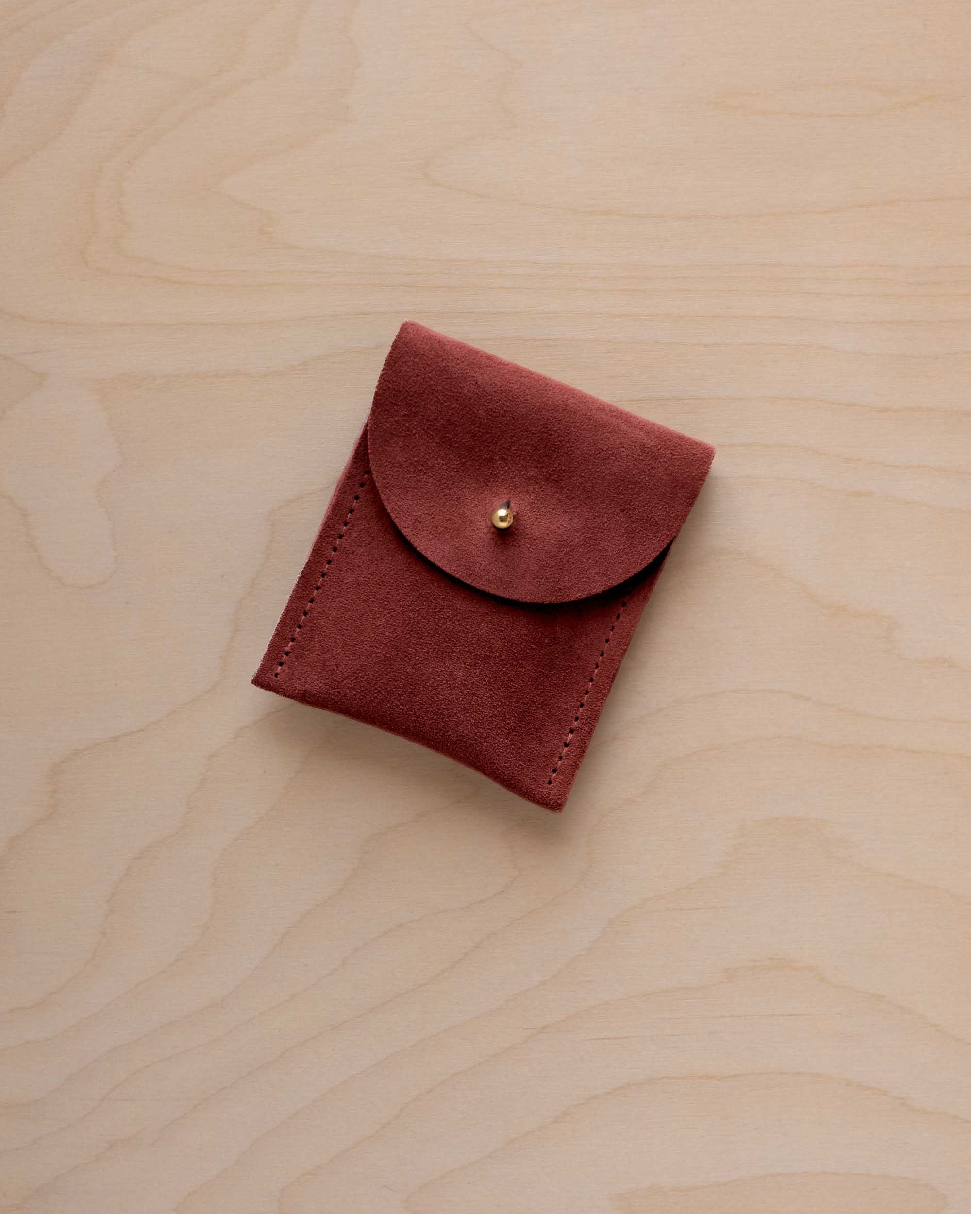A small rust red coloured suede pouch with a gold coloured fastening and flap closure, placed on a light wood grain surface.