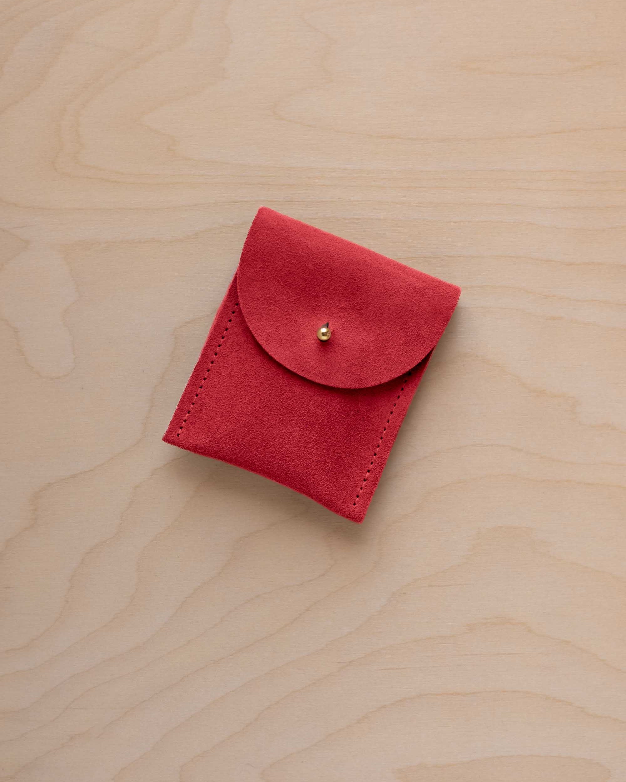 A small scarlet red coloured suede pouch with a gold coloured fastening and flap closure, placed on a light wood grain surface.