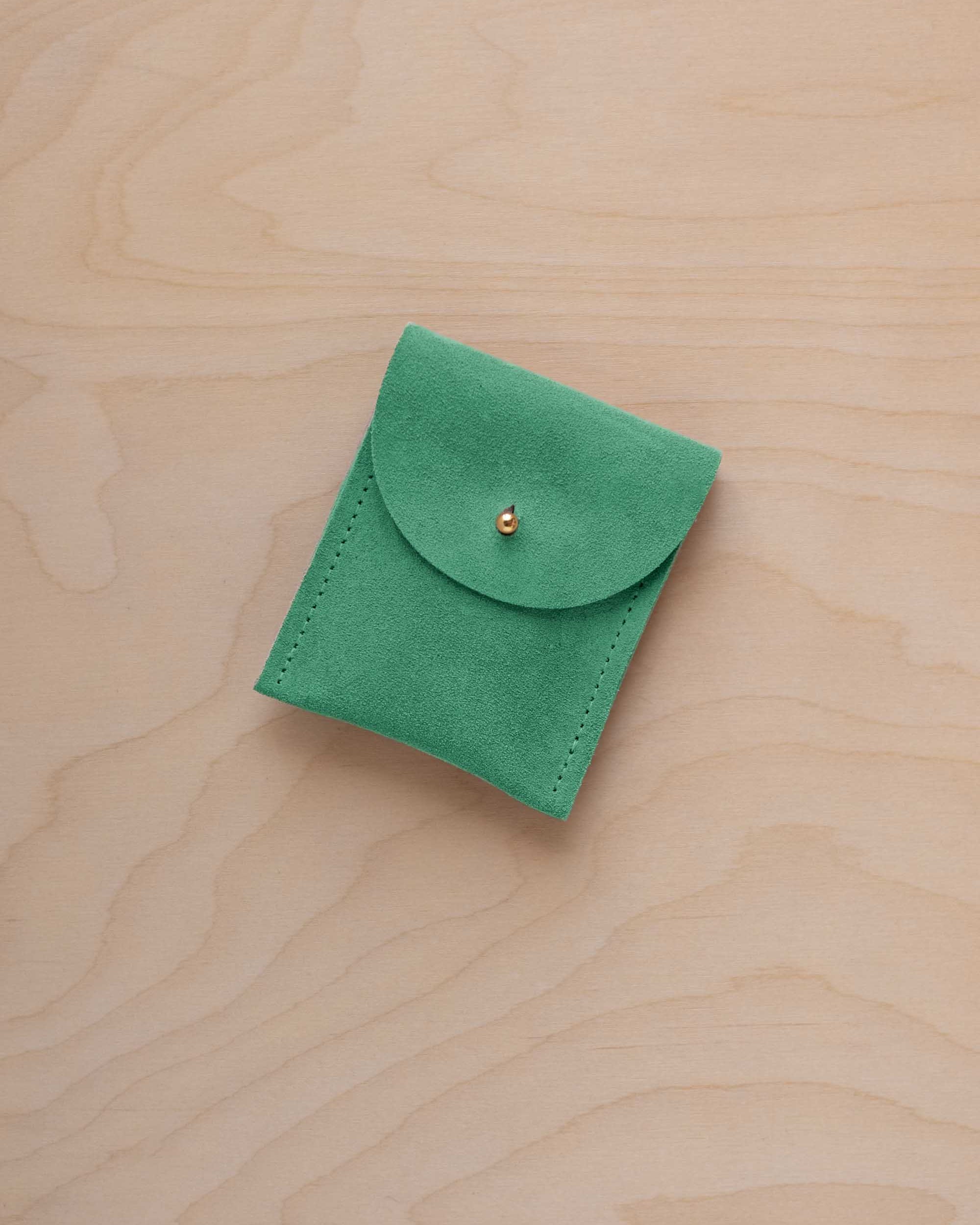 A small seafoam green coloured suede pouch with a gold coloured fastening and flap closure, placed on a light wood grain surface.