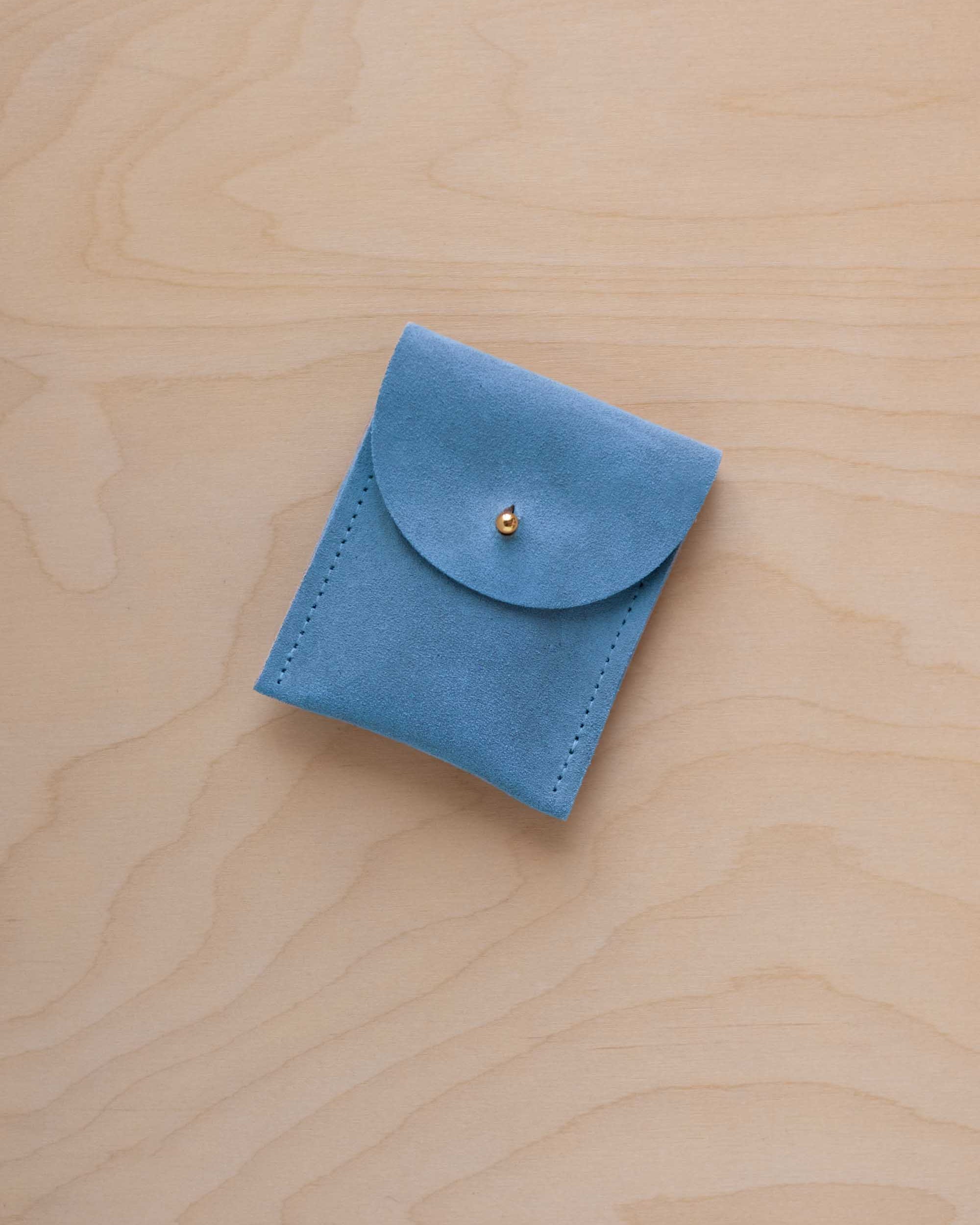 A small sky blue coloured suede pouch with a gold coloured fastening and flap closure, placed on a light wood grain surface.