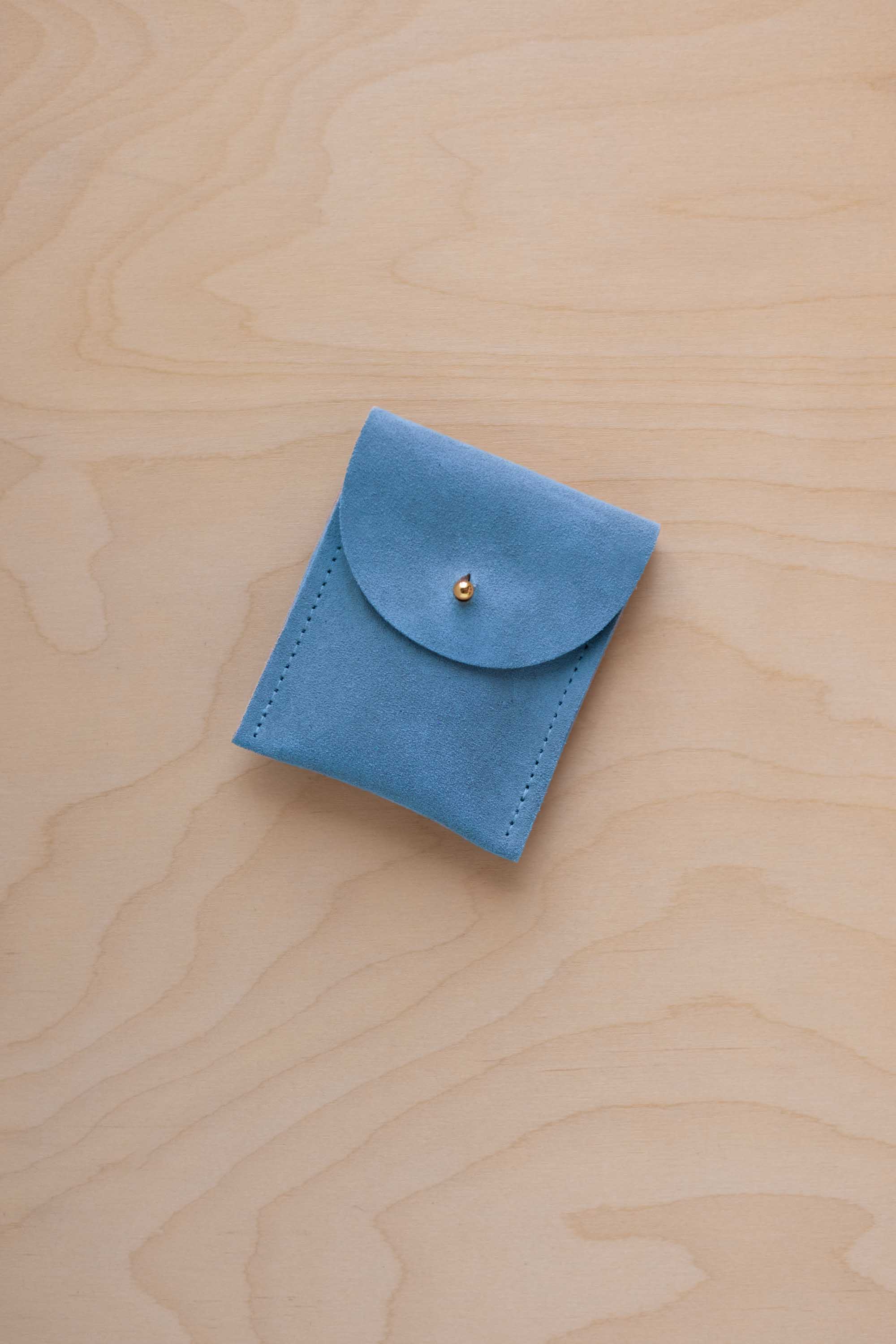 A small sky blue coloured suede pouch with a gold coloured fastening and flap closure, placed on a light wood grain surface.