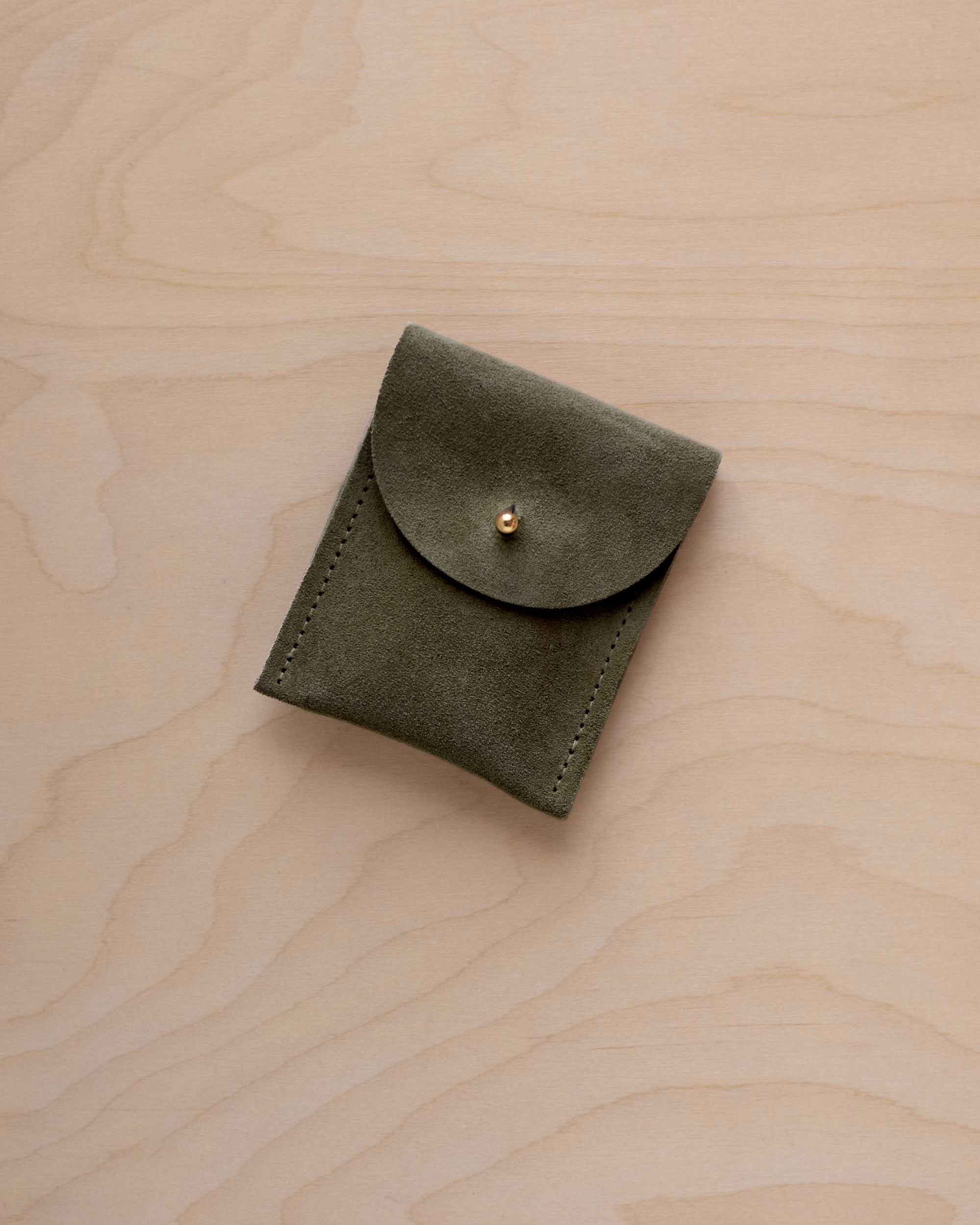 A small soft olive green coloured suede pouch with a gold coloured fastening and flap closure, placed on a light wood grain surface.