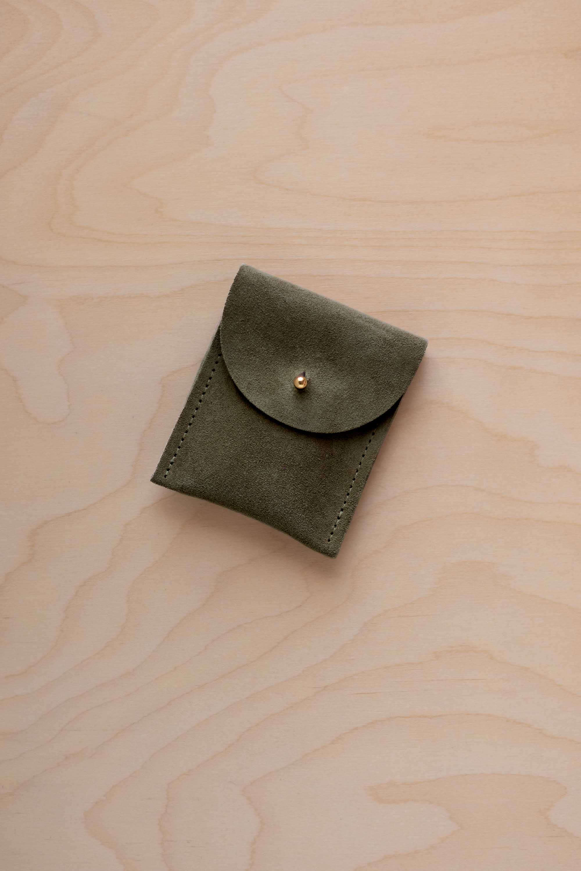 A small soft olive green coloured suede pouch with a gold coloured fastening and flap closure, placed on a light wood grain surface.