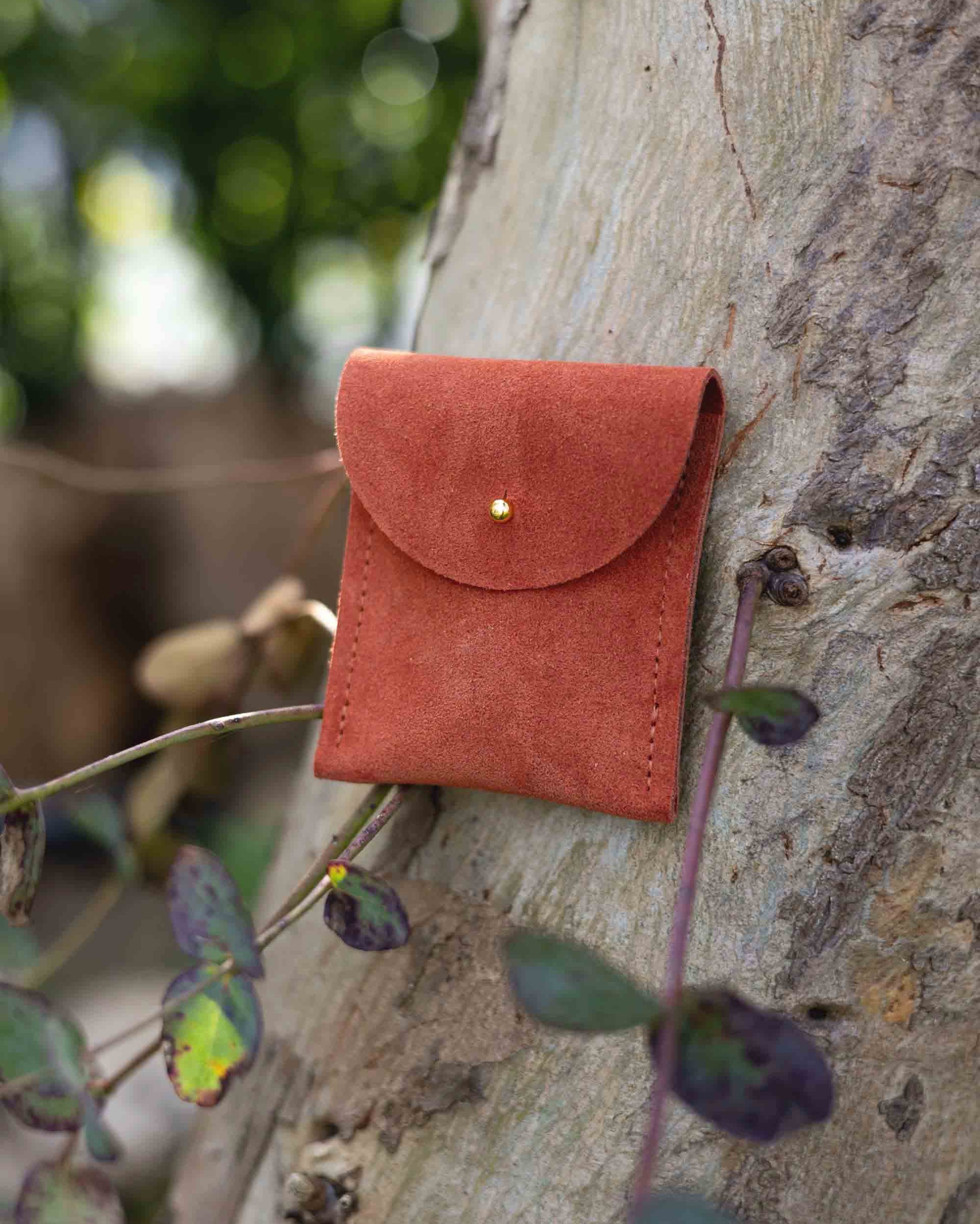 A small terracotta coloured suede pouch with a gold coloured fastening and flap closure, leaning against a natural wooden surface with an outdoor background.