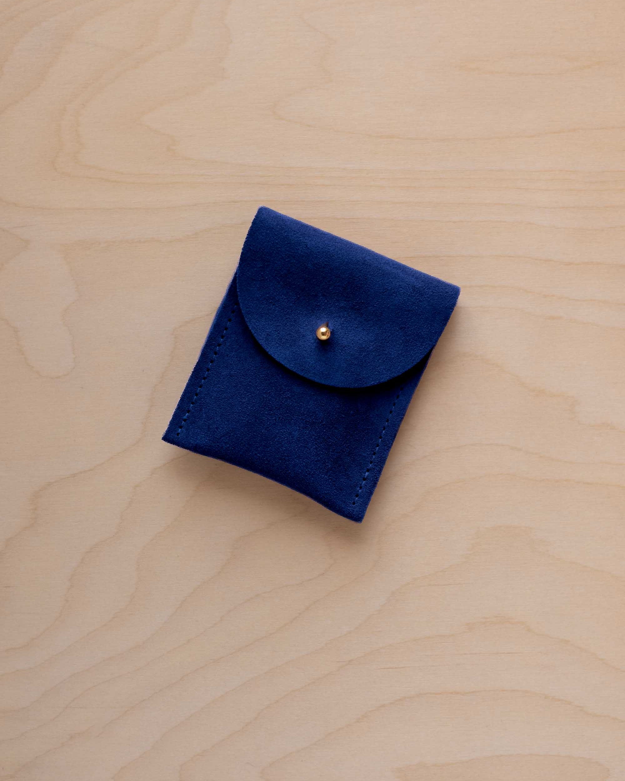 A small ultramarine blue coloured suede pouch with a gold coloured fastening and flap closure, placed on a light wood grain surface.