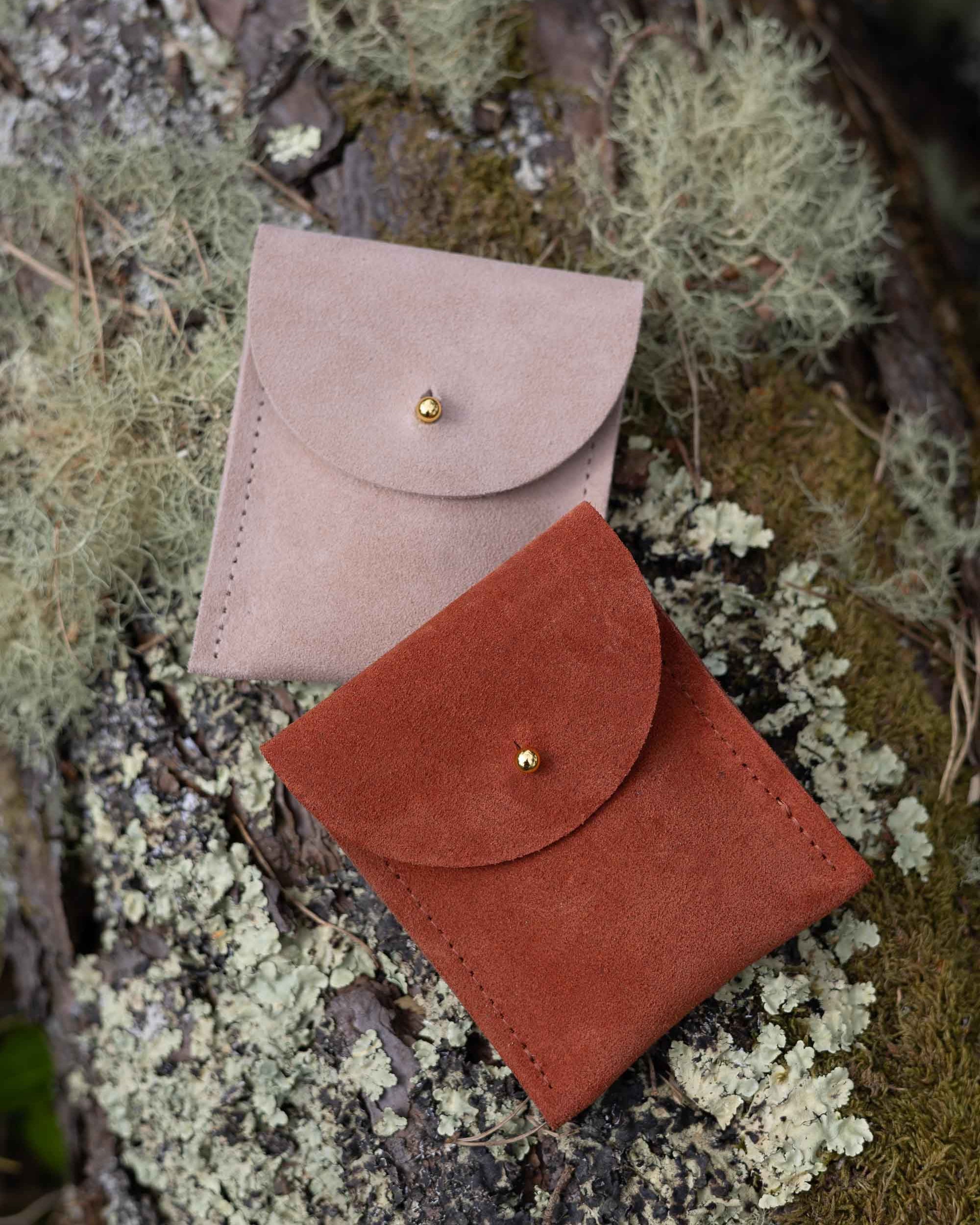 Two small coloured suede pouches with a flap closure and a gold coloured fastening, on a natural outdoor background.
