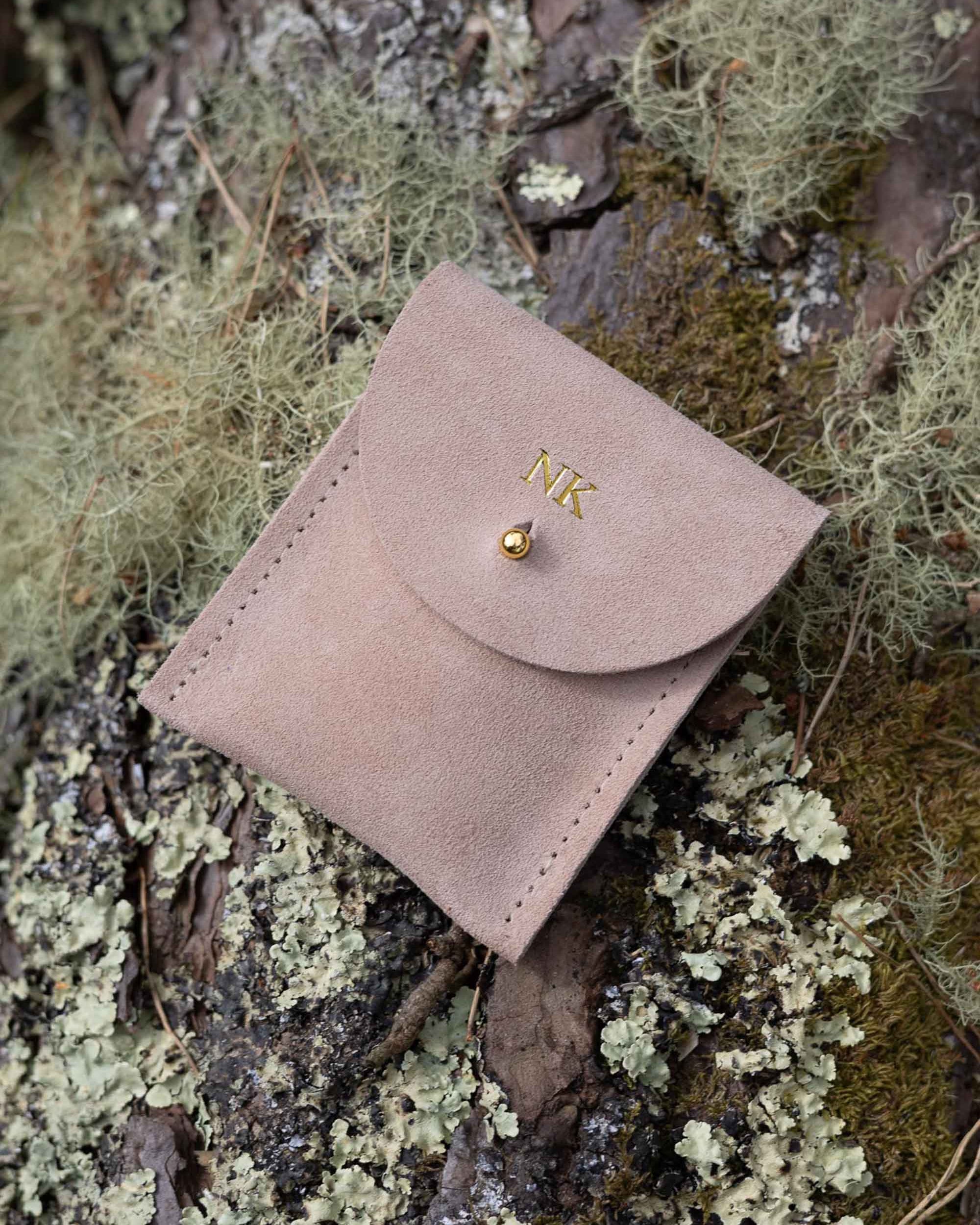 A top-down view of a warm sand coloured suede pouch with a flap closure and a gold coloured fastening, on a natural outdoor background.