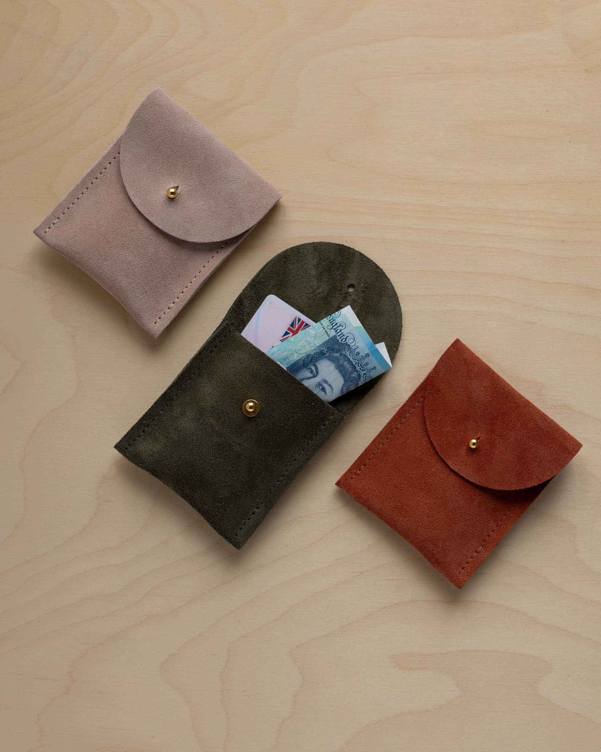 Three small suede pouches in assorted colours with a flap closure and a gold coloured fastening, arranged in a row on a light wood grain surface. The middle pouch has a bank card and a bank note nestled inside.