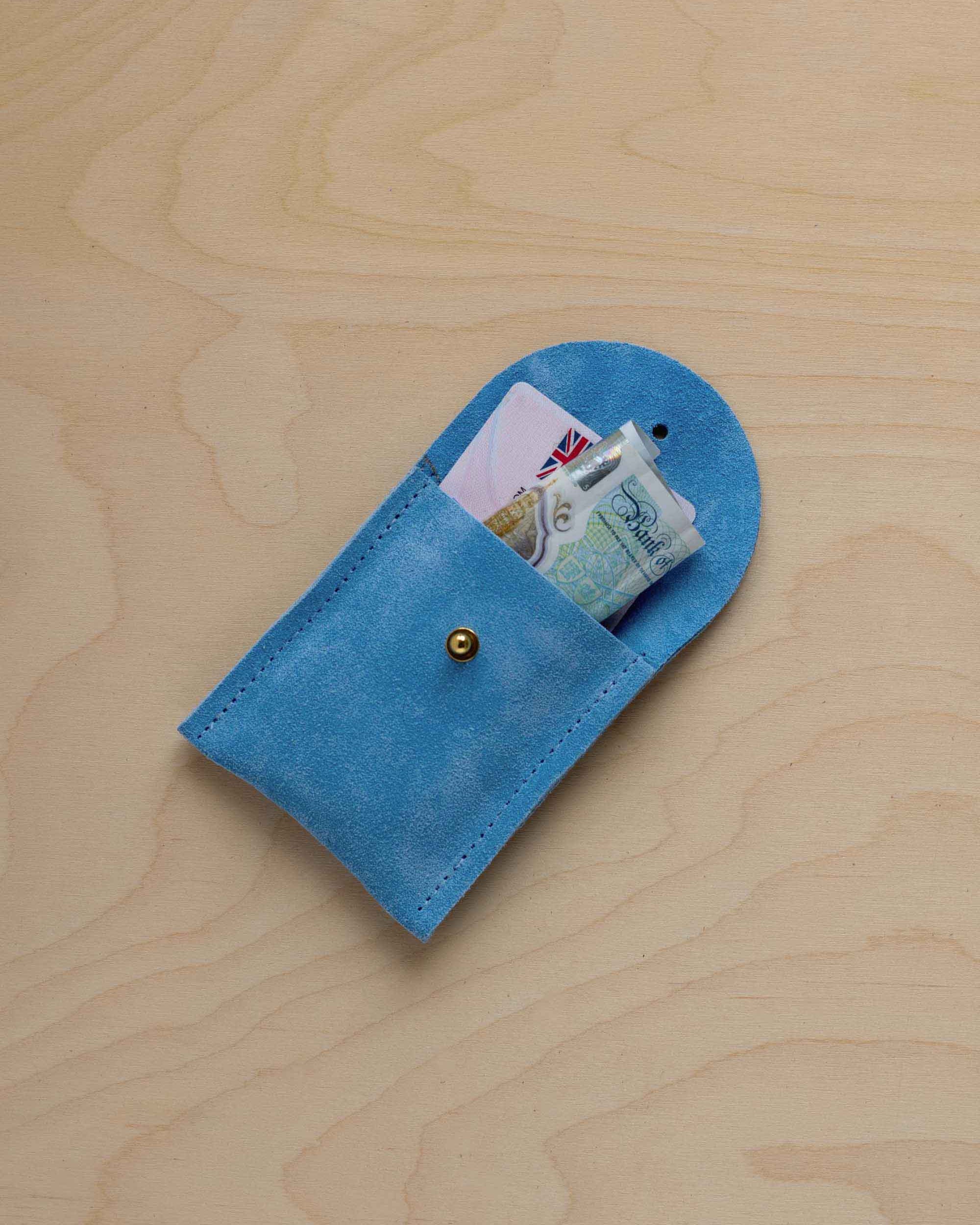 A small sky blue coloured suede pouch with a gold coloured fastening and flap closure, placed on a light wood grain surface, with a bank card and a bank note nestled inside.