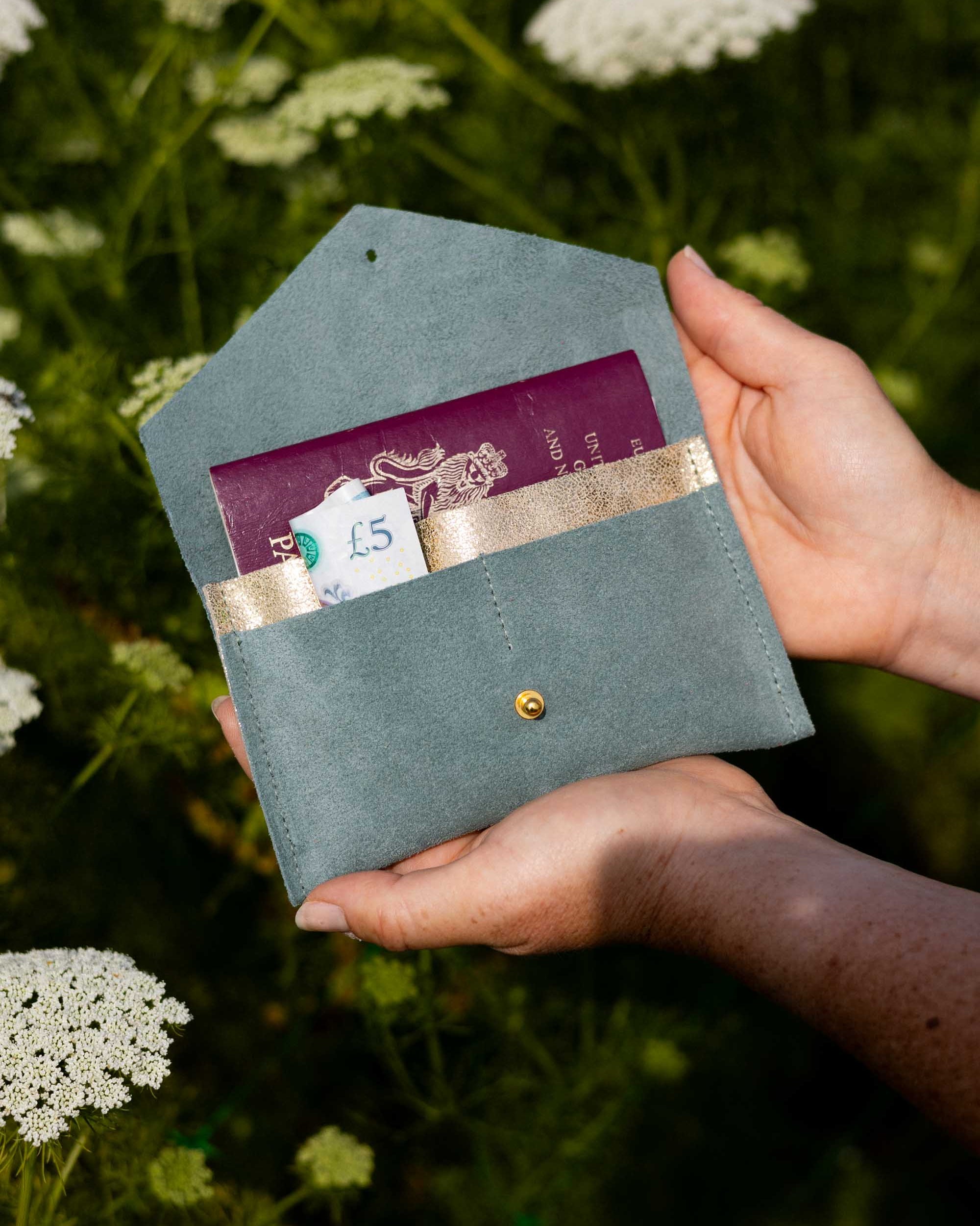 A person's hands holding an open eucalyptus or sage green envelope-style wallet with a gold-tone button, showing a purple passport and cards inside, set against a blurred background of green foliage and white wildflowers.