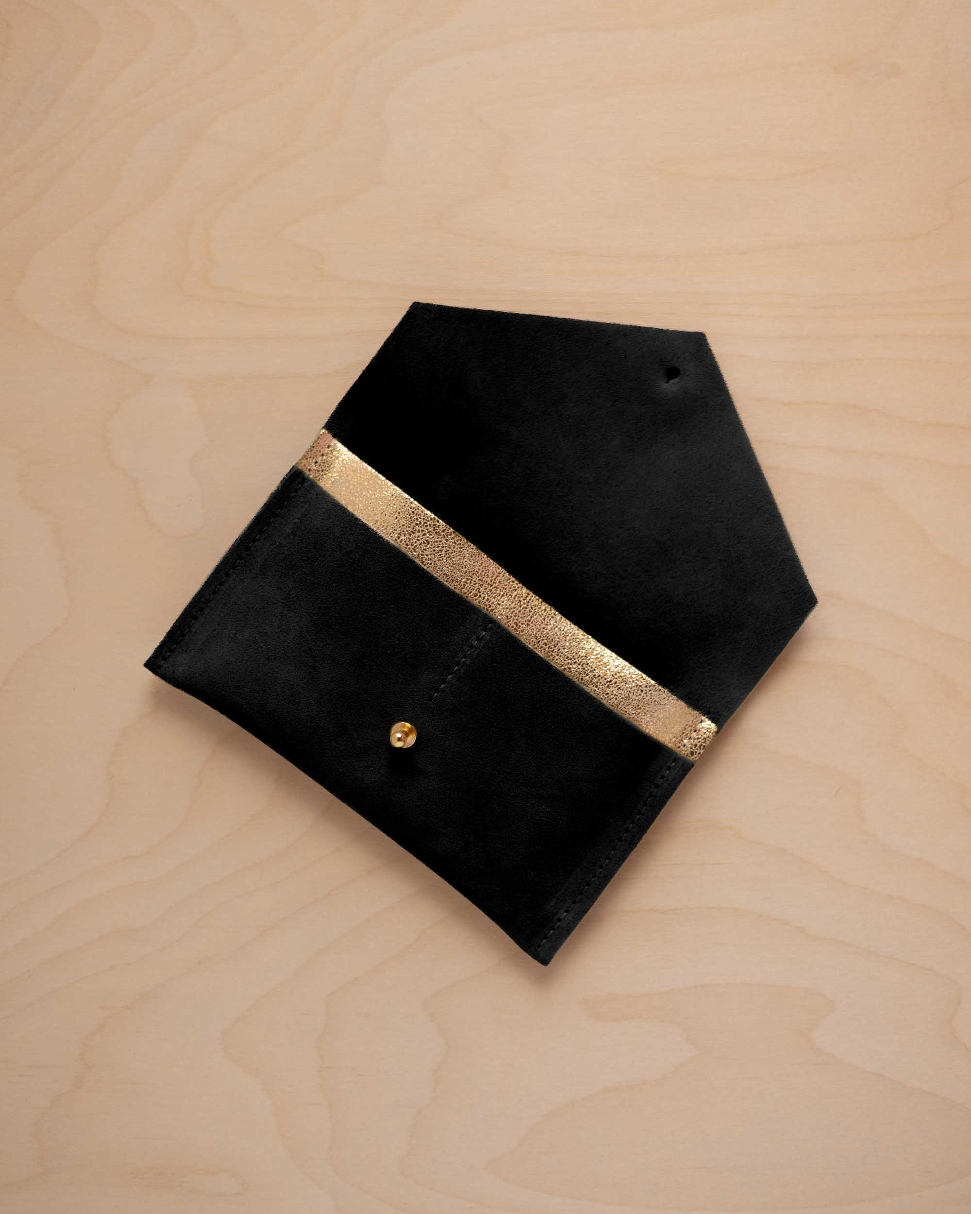 A black suede pouch with a metallic gold contrast pocket and a small gold stud, displayed on a light wood grain background.