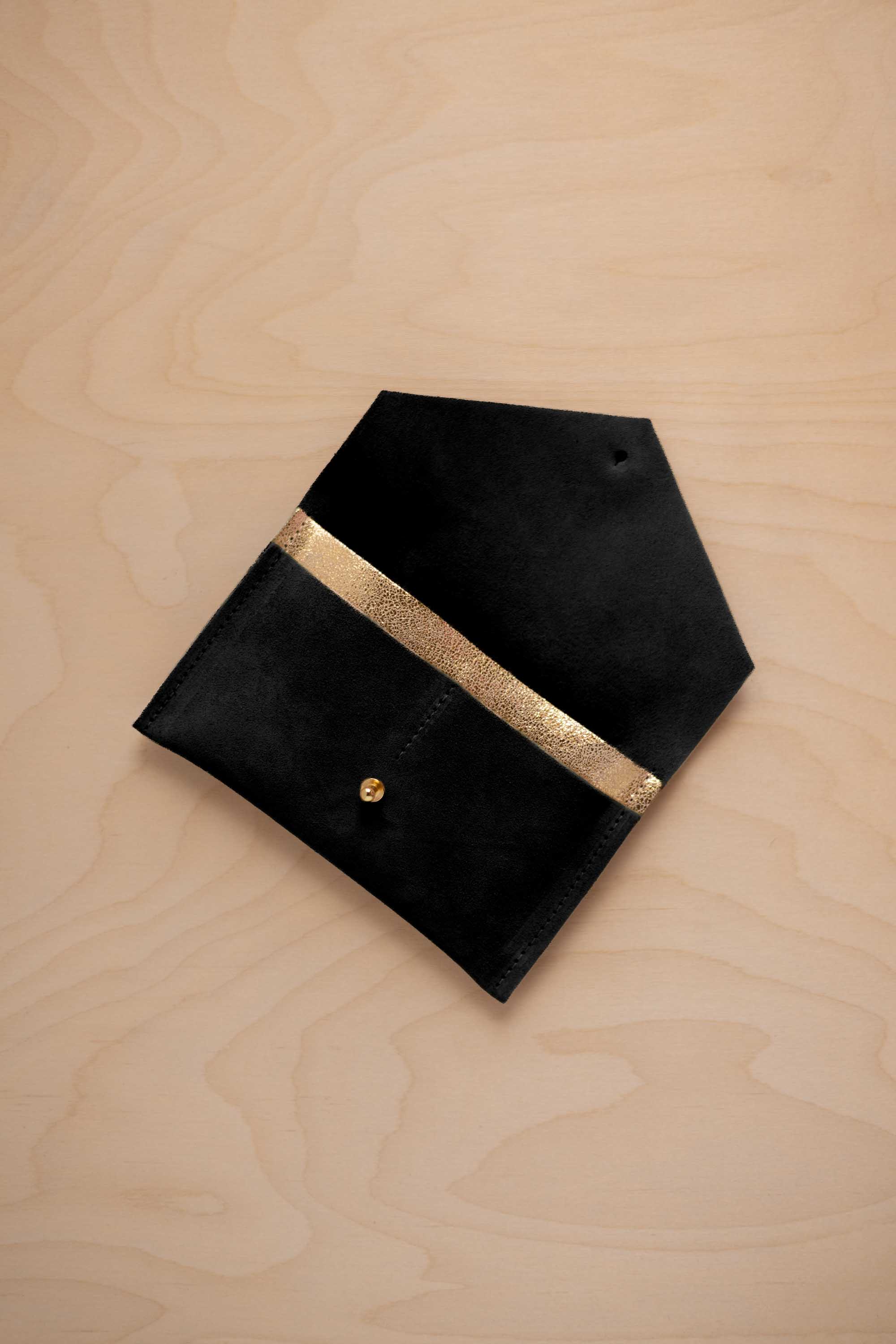 A black suede pouch with a metallic gold contrast pocket and a small gold stud, displayed on a light wood grain background.